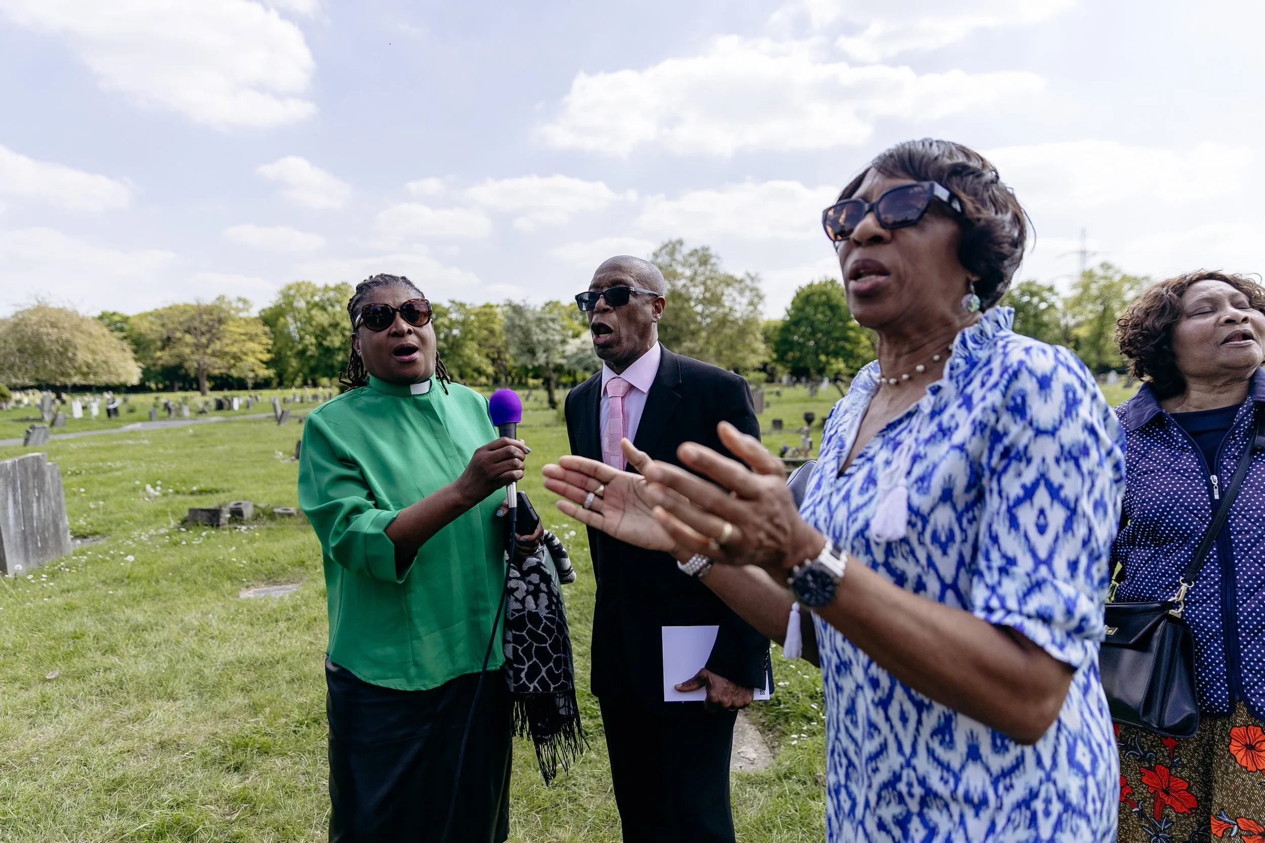 Group of four people, two women and two men, standing outdoors, engaging in conversation; one woman is wearing glasses and a blue and white patterned blouse, another woman is holding a microphone and wearing a green clergy robe, a man in a suit appea