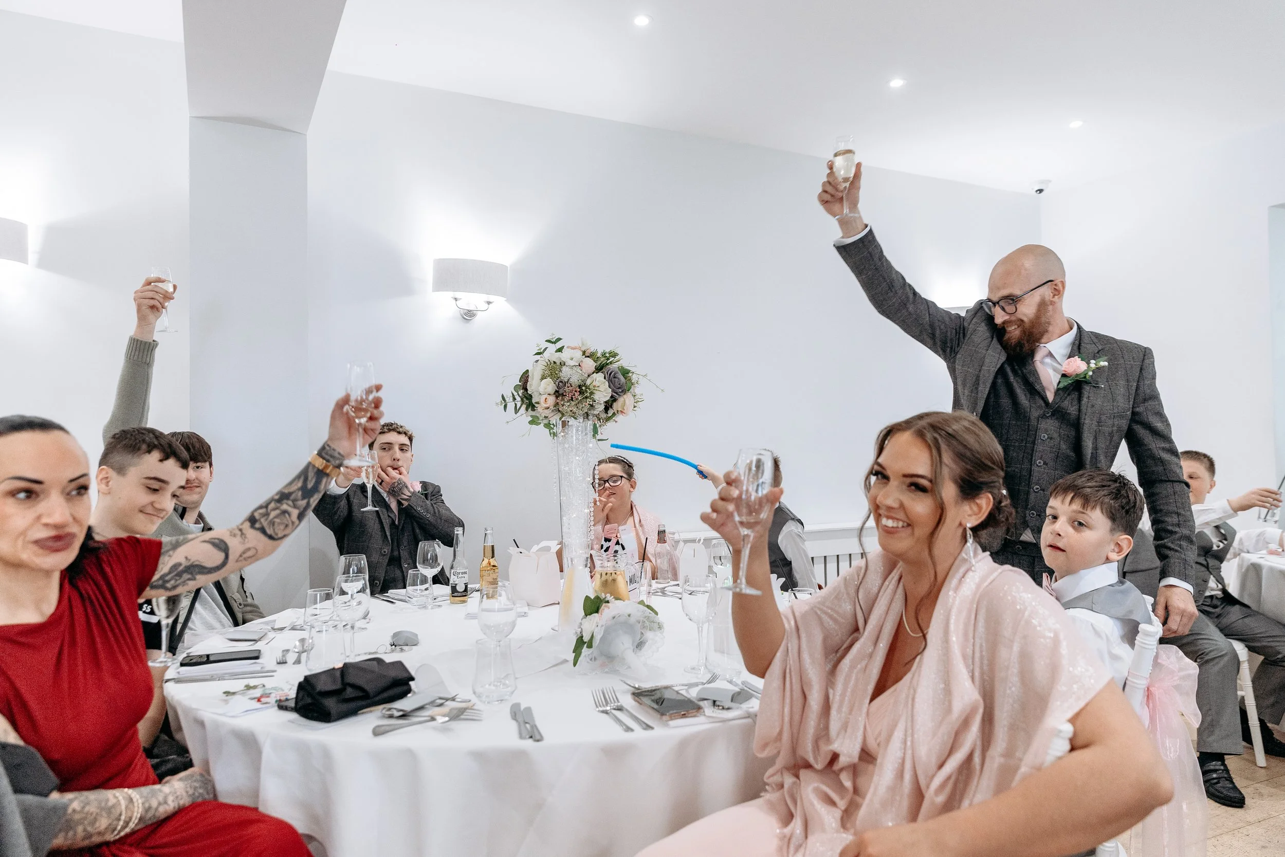 People celebrating at a wedding reception, raising glasses in toast, with a man standing and smiling among seated guests at a decorated table.