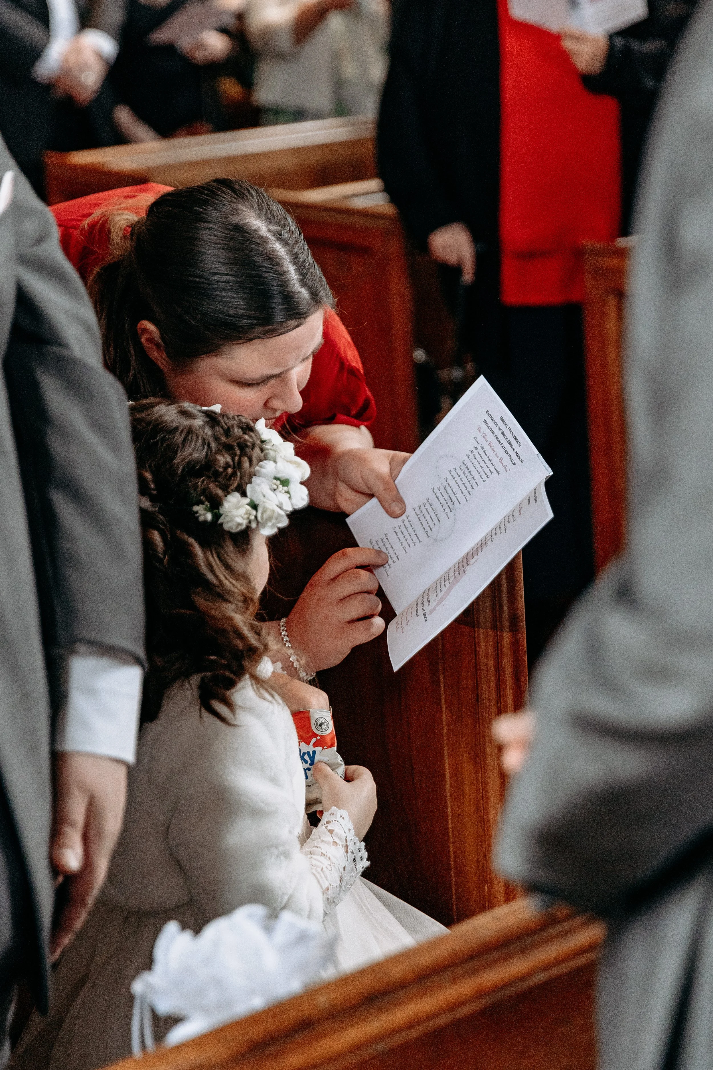 A woman and a young girl, likely at a religious ceremony, are reading a booklet together in a church. The girl is holding a snack, wears a white dress with lace sleeves and a floral headband. The woman has dark hair and is dressed in red.