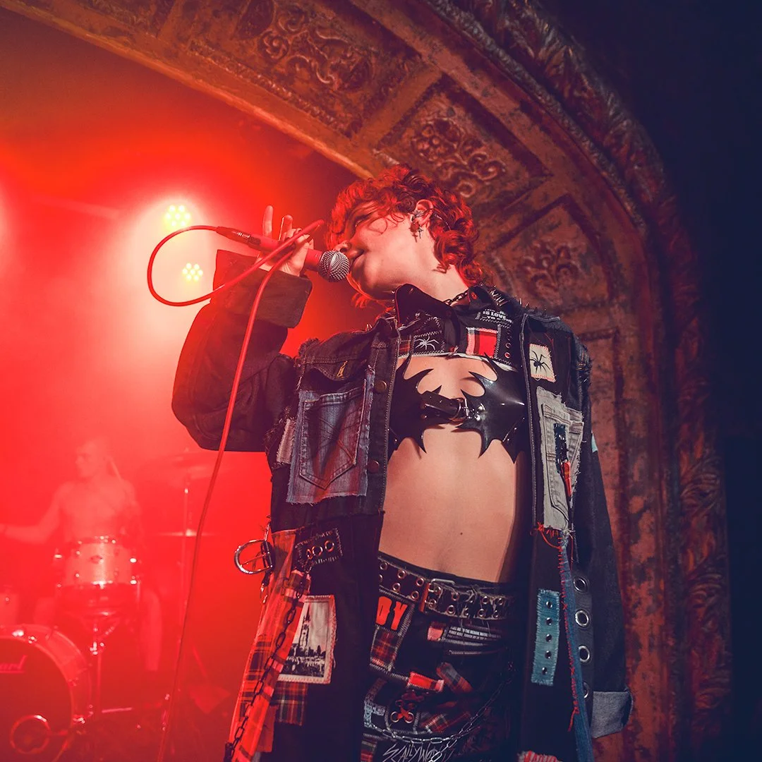 A singer with curly red hair performing with a microphone on stage, dressed in a black leather top and revealing clothing, under red stage lighting with ornate decorations in the background.