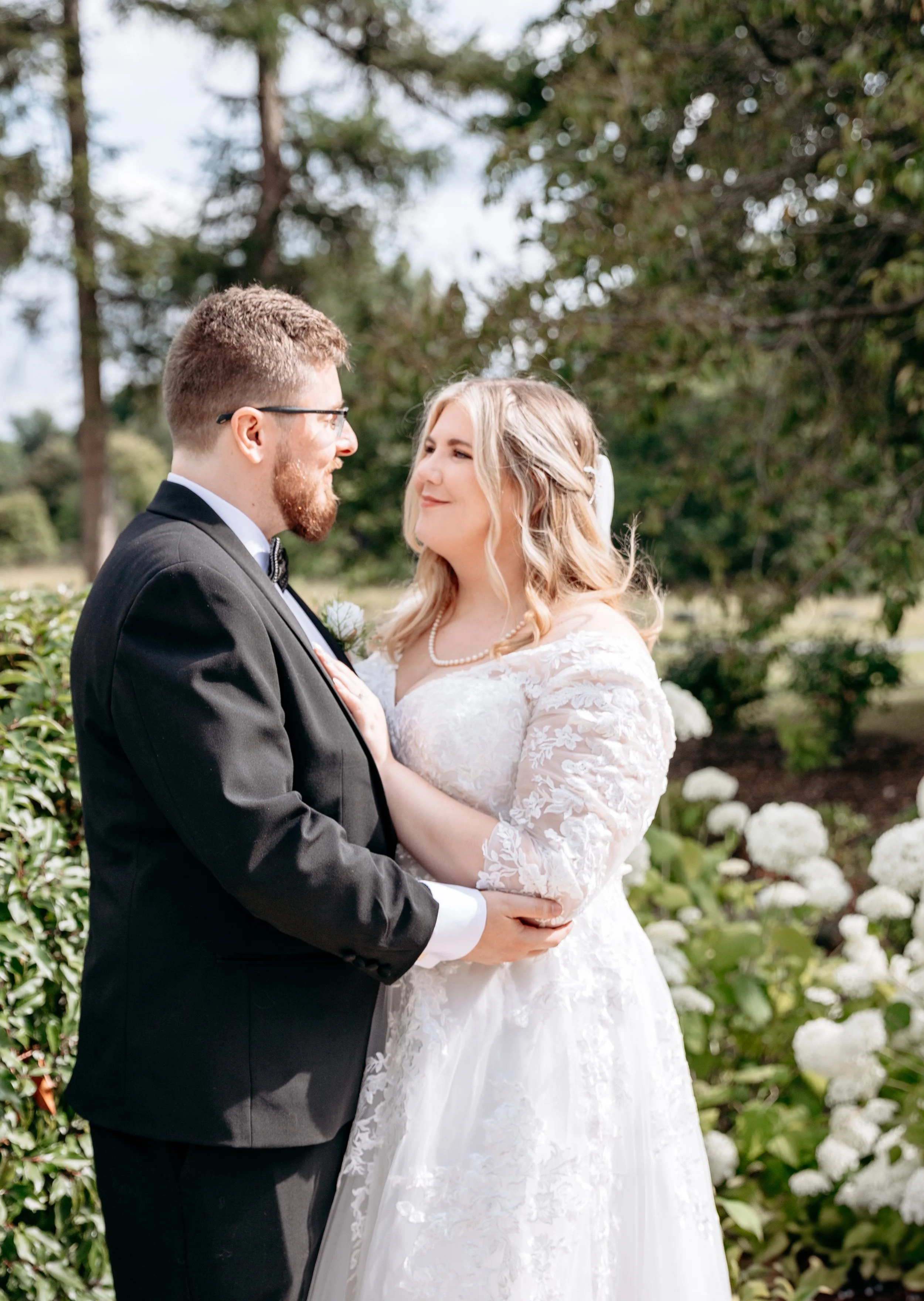 Bride and groom embracing in a garden setting, with the bride in a white lace gown and the groom in a black suit.