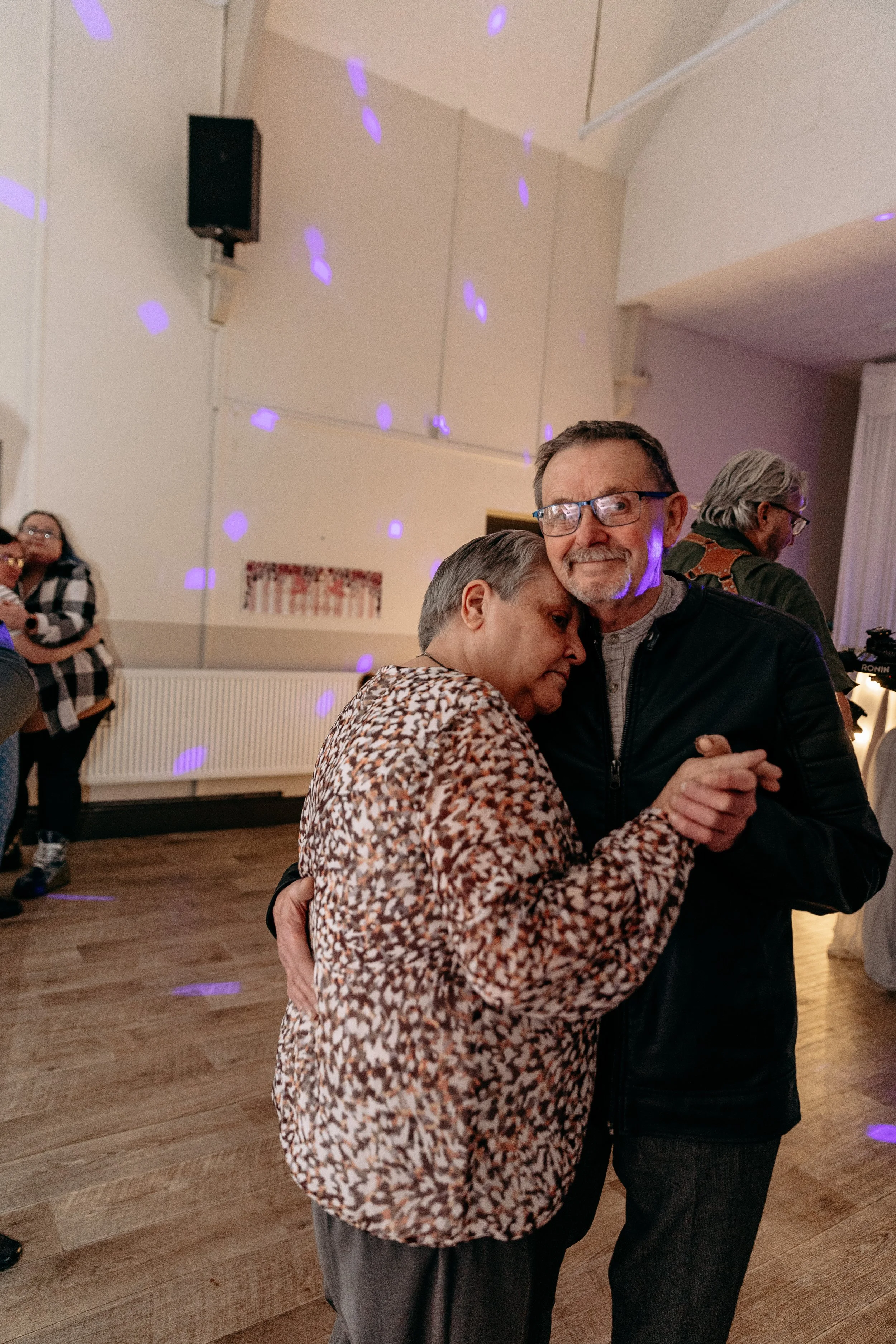 A couple dancing closely on a wooden floor in a social setting with other people in the background, purple light spots on the wall.