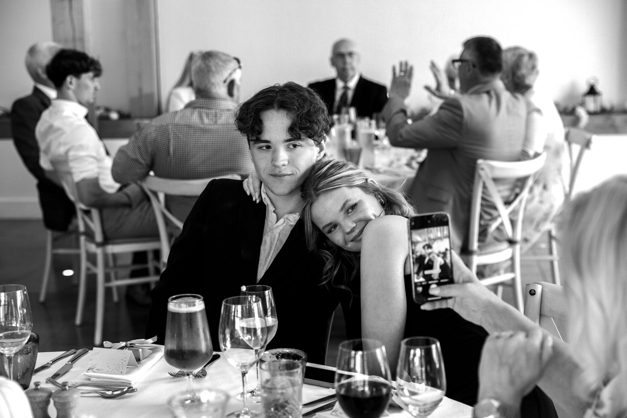 A black and white photo of a young couple sitting together at a restaurant table. The woman leans on the man's shoulder while holding a smartphone to take a picture. In the background, other people are seated at tables, engaged in conversation.