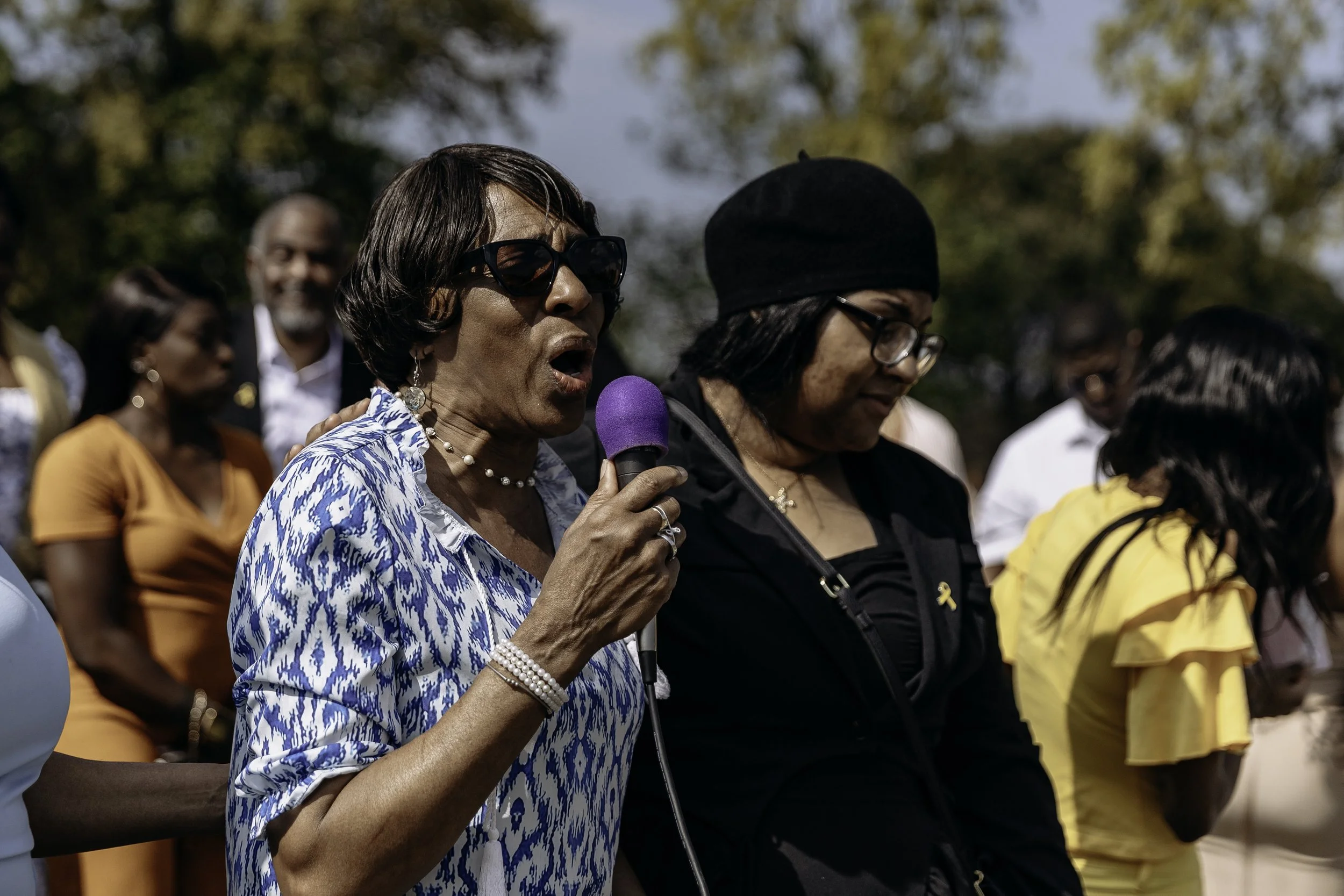 Woman wearing sunglasses and a blue patterned shirt speaking into a purple microphone at a gathering outdoors.