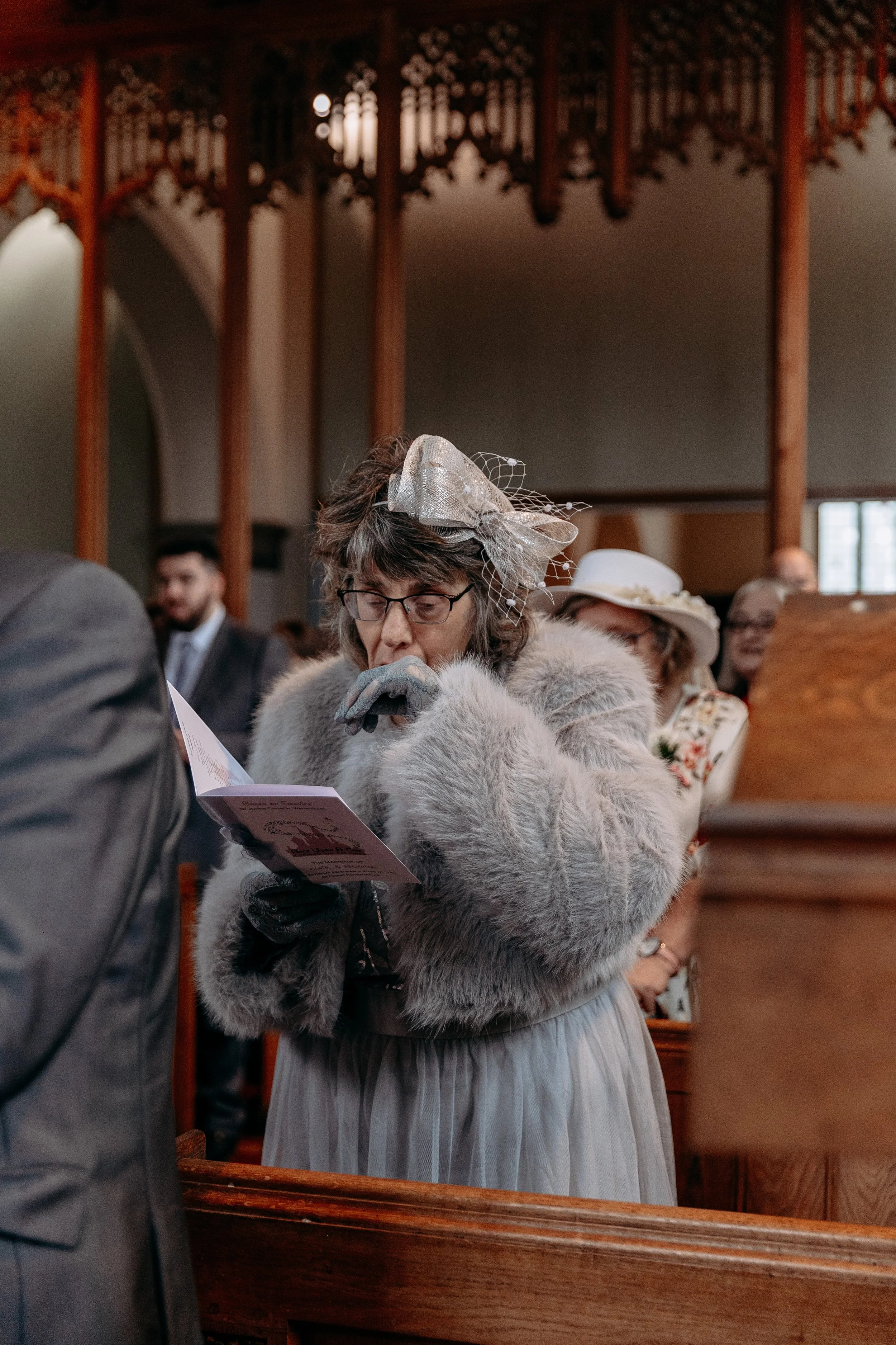 A woman dressed in vintage attire with a large decorative bow on her head, reading a program or booklet at a formal event inside a church or chapel with wood paneling.