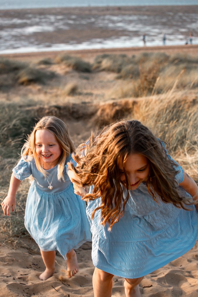Two girls in blue dresses running up sandy dunes near the beach.