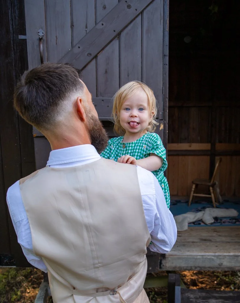 A man with short hair and a beard holding a young girl with blonde hair and blue eyes, wearing a green and white checkered dress, outside near a wooden shed or barn, with a small chair inside.