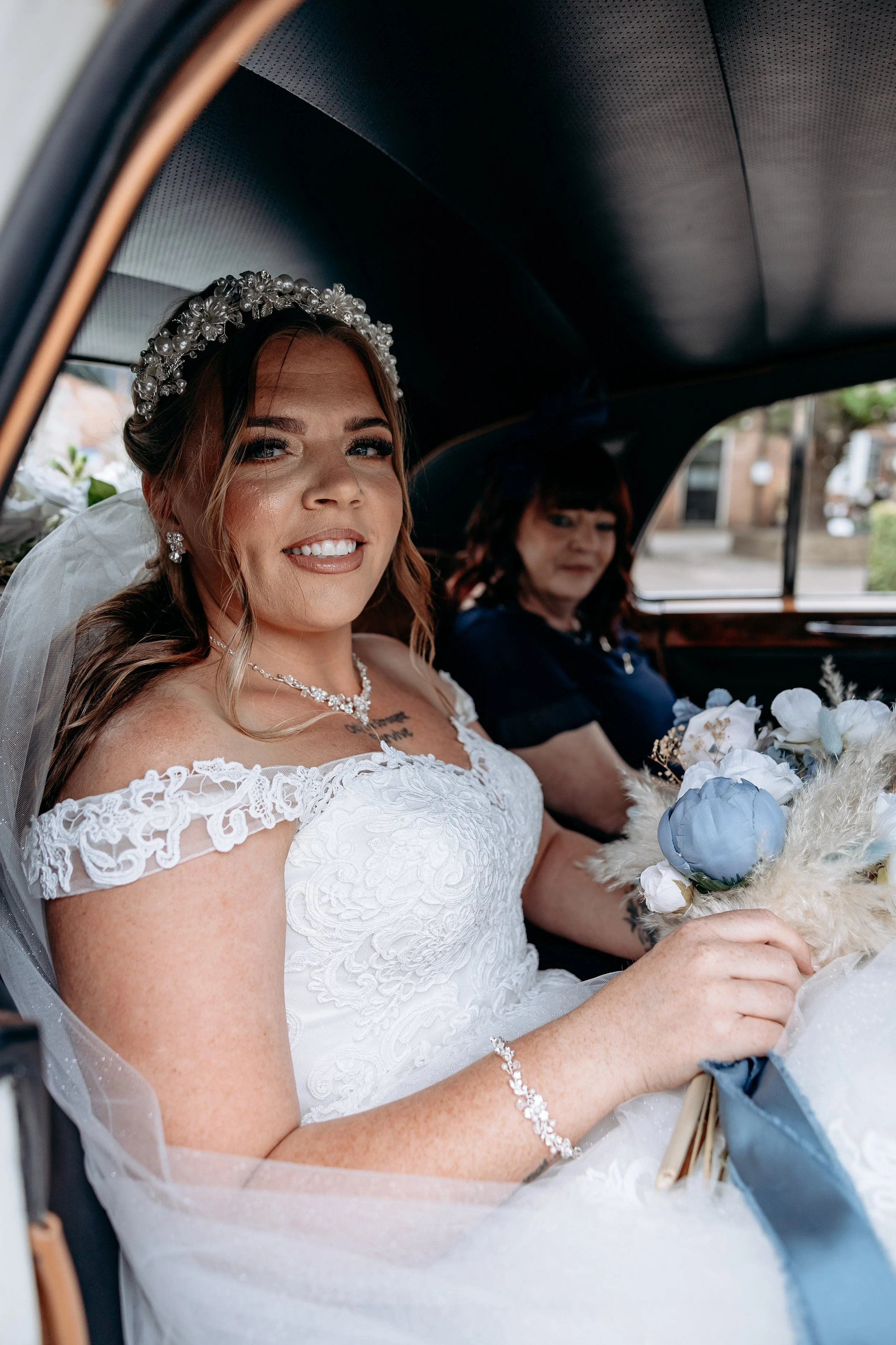 A bride sitting in a car, smiling, holding a bouquet of white and blue flowers, wearing a white lace wedding dress, with a pearl headband and jewelry, accompanied by a woman in a dark outfit.