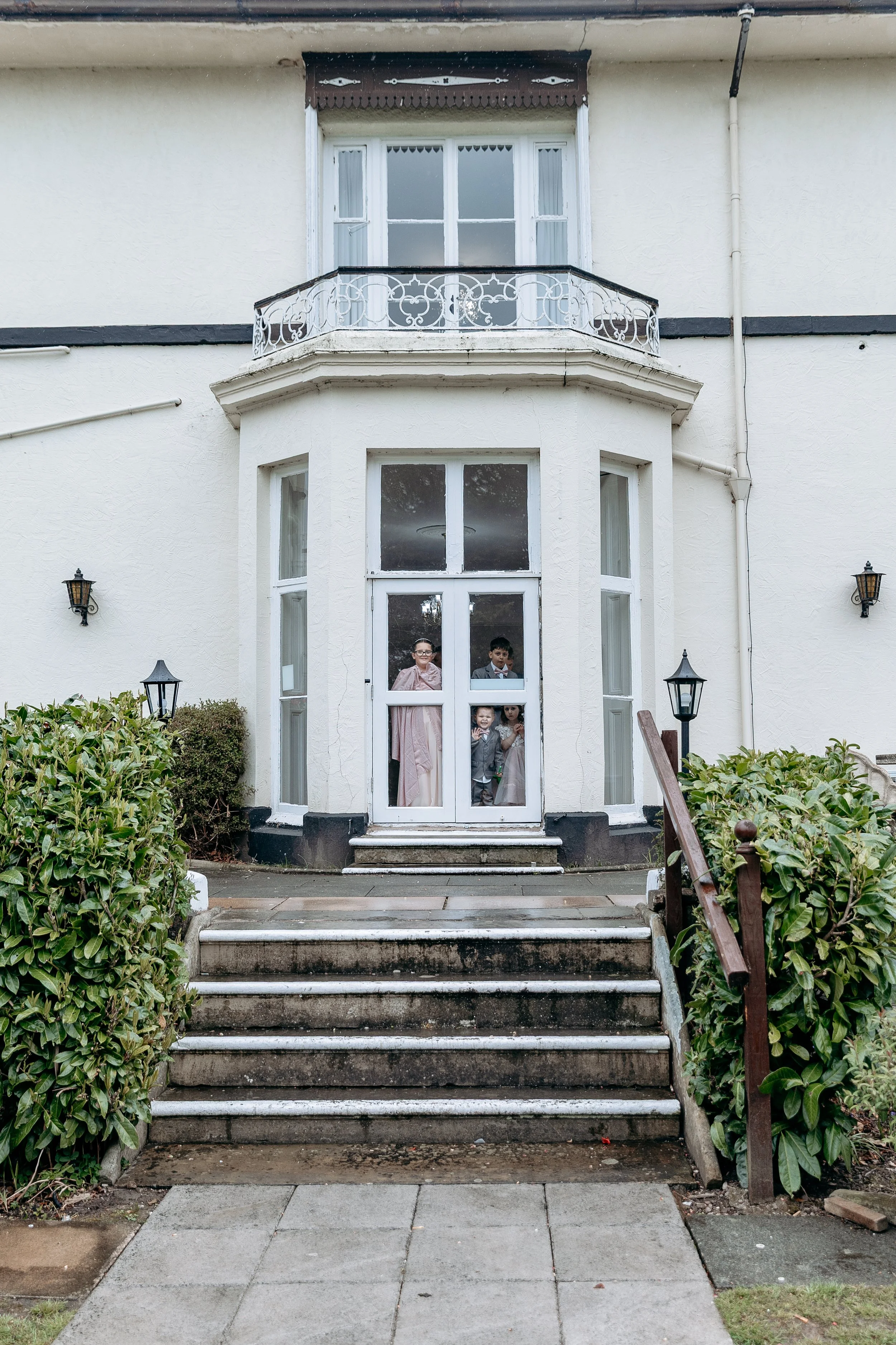 Children looking through the front door of a house with stairs, bushes, and lanterns outside.
