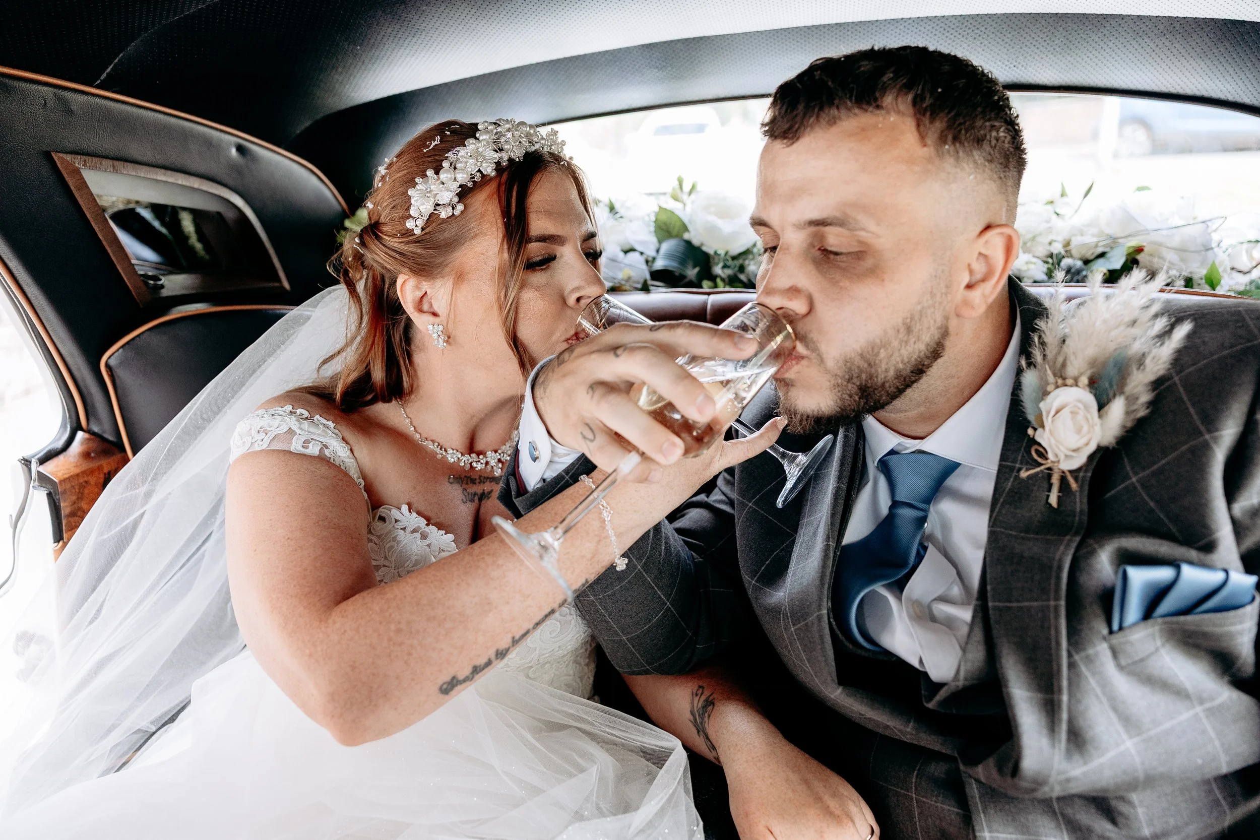 Bride and groom in wedding attire sharing a drink in the back of a vintage car decorated with flowers.