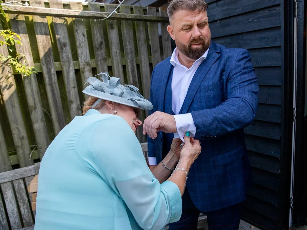 A woman wearing a light blue outfit and a large gray hat is pinning a badge onto a man in a blue checkered suit jacket in a backyard with a wooden fence.