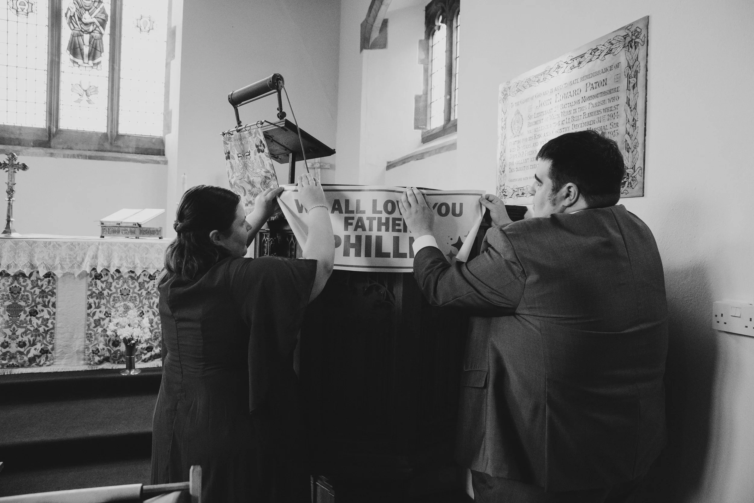 Two people are hanging a banner that reads 'WE ALL LOVE YOU FATHER PHIL' in a church.