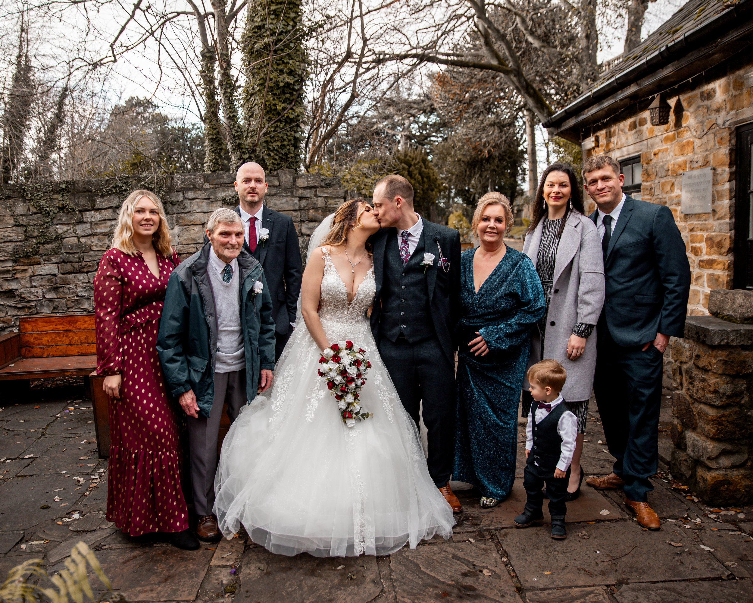 A group of people celebrating a wedding outdoors, with the bride and groom kissing in the center. The bride is in a white wedding dress holding a bouquet, and the groom is in a dark suit. Family and friends surround them against a stone wall and rust