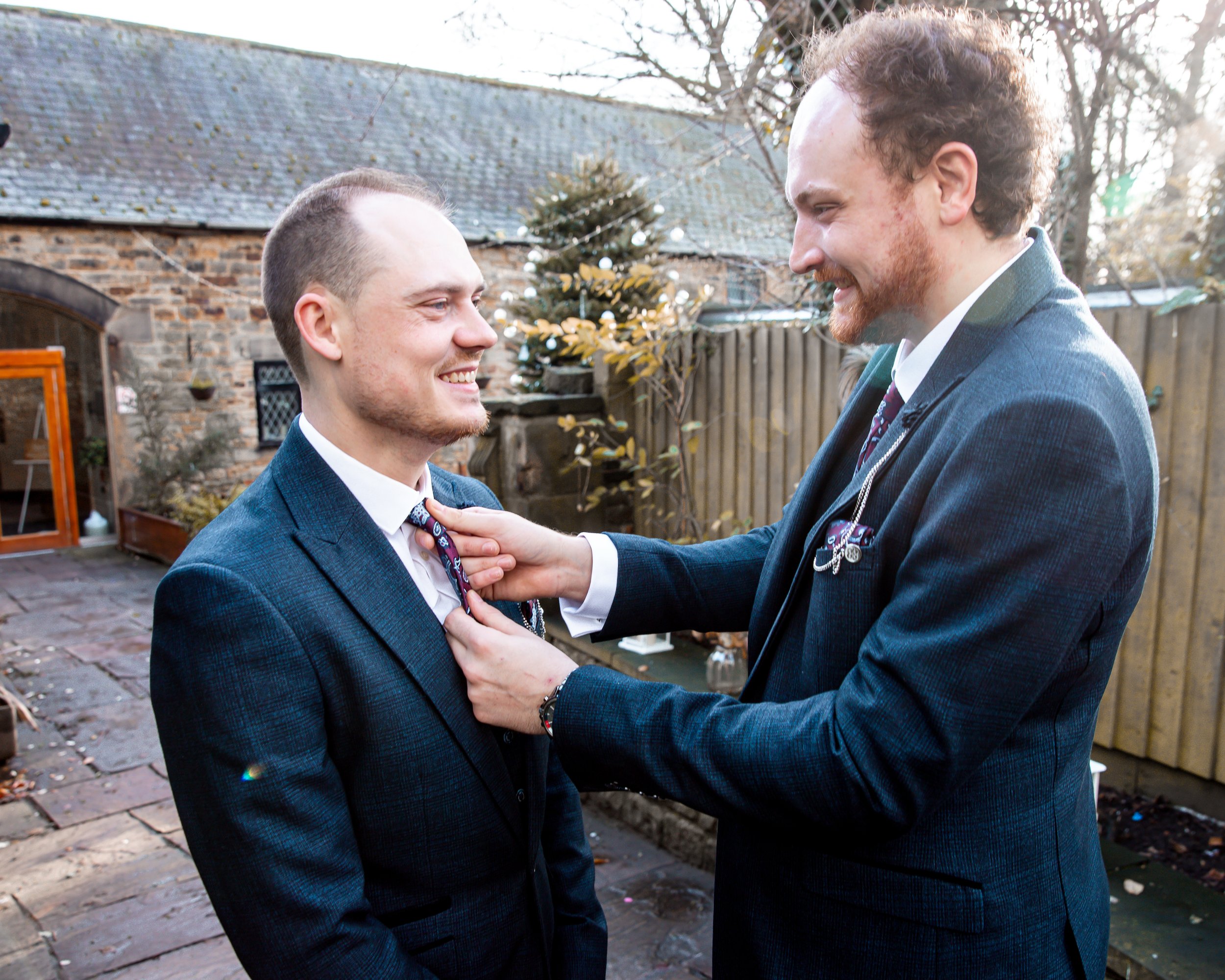 Two men in suits smiling and one adjusts the other's tie outdoors on a paved patio with a brick building, wooden fence, and trees in the background.