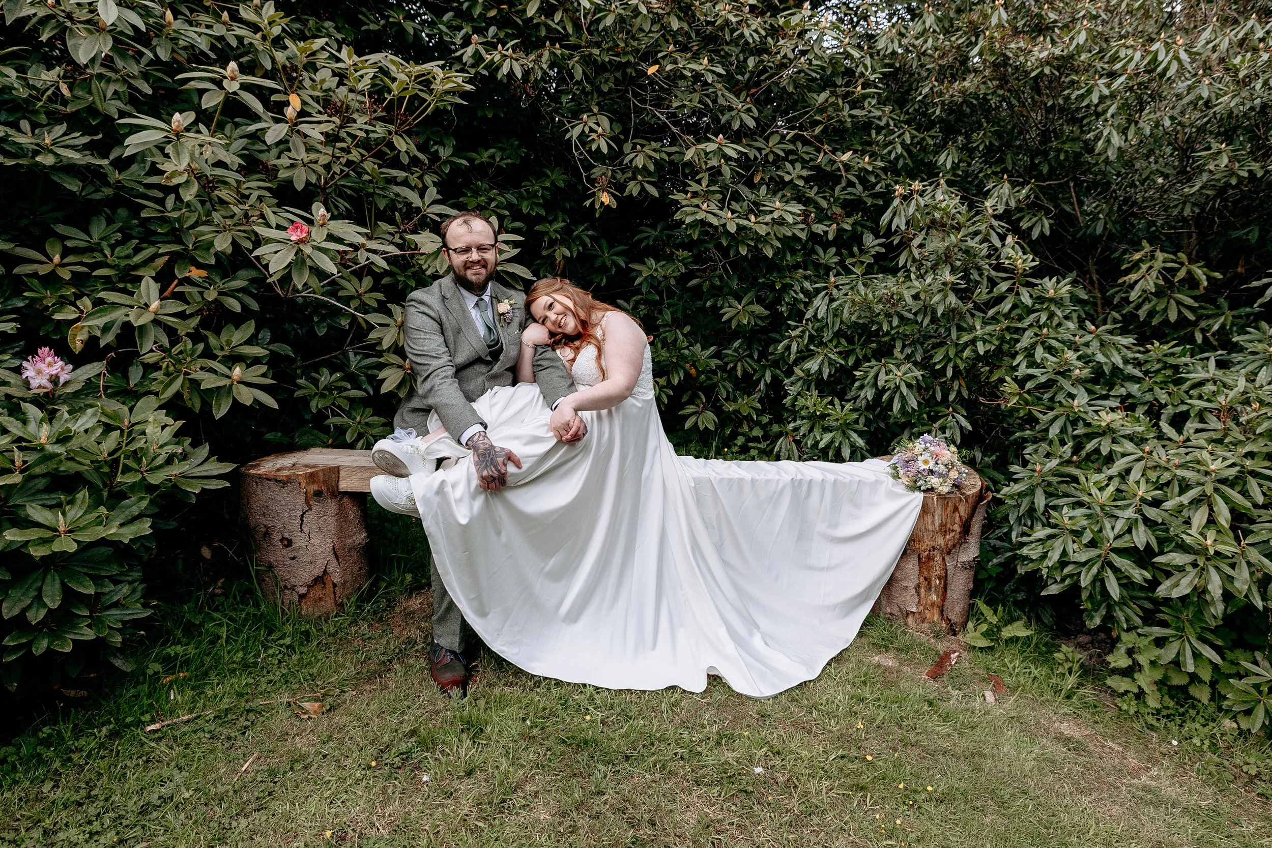 A couple in wedding attire sitting on a wooden log bench in front of green foliage, smiling and enjoying their special day. The bride is wearing a white dress with a train and the groom is in a gray suit with tattoos on his arm.