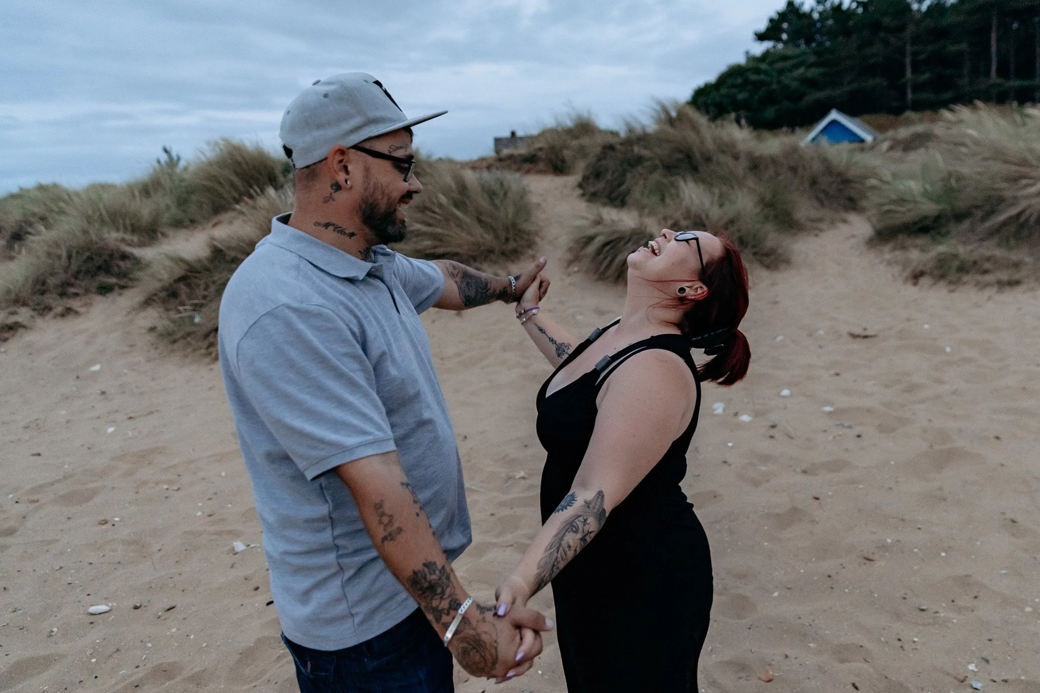 A couple holding hands and smiling on a sandy beach with dunes and grasses in the background during cloudy weather.