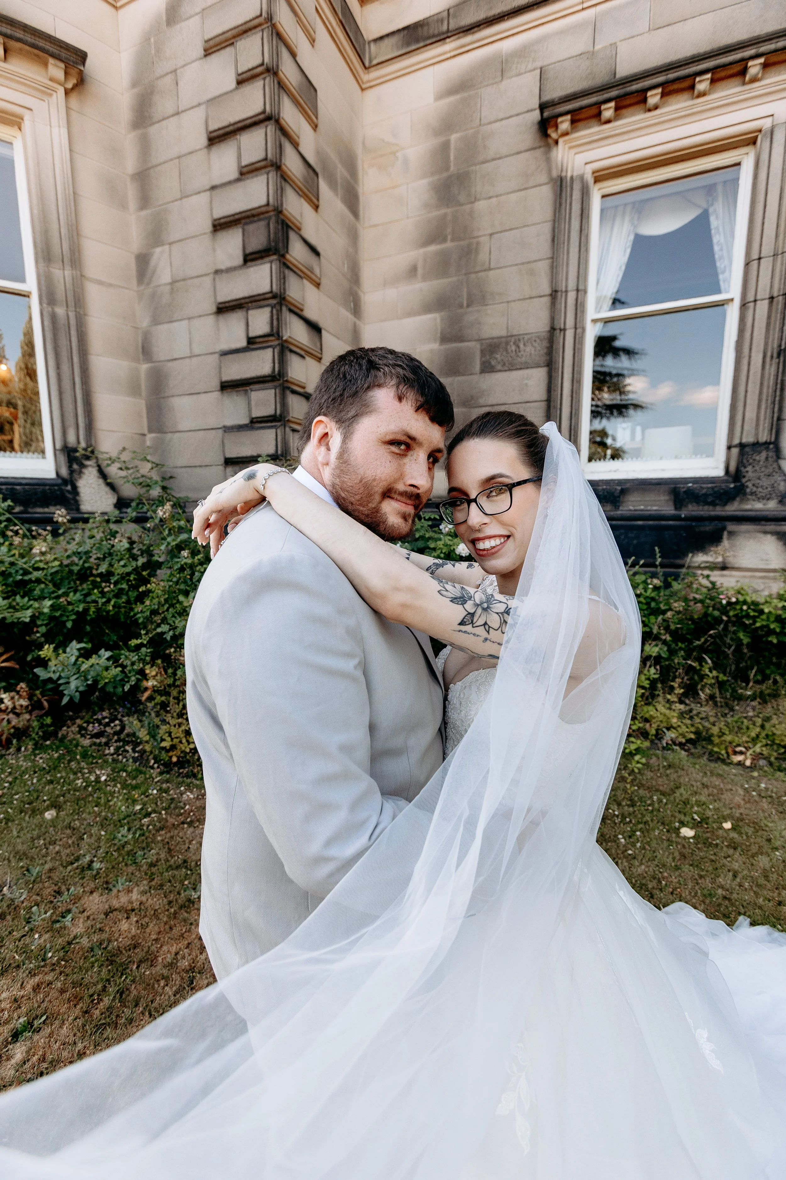 A bride and groom pose together outdoors in front of a stone building, both smiling and embracing.