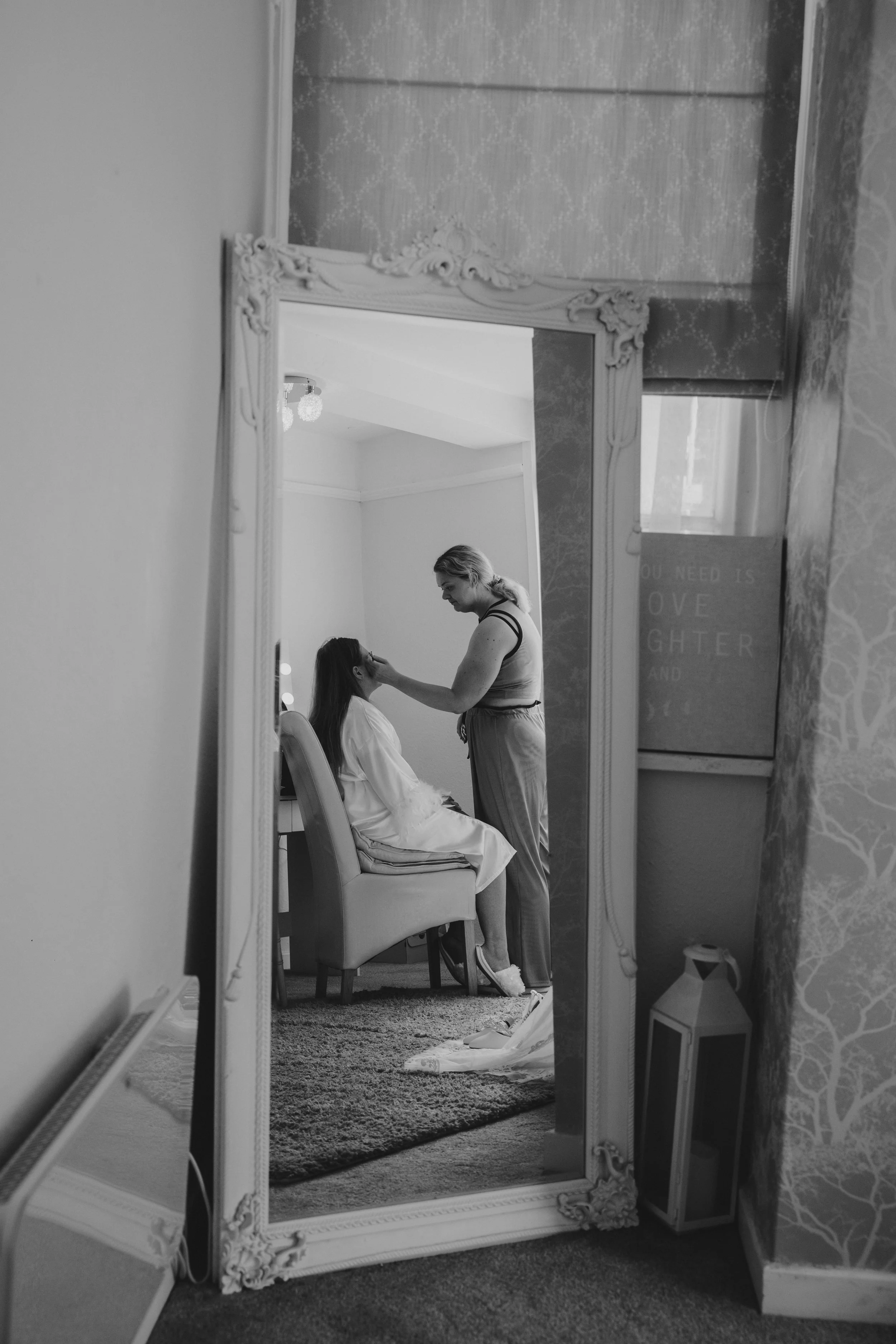 A woman getting her makeup done in a bedroom, seen through a reflection in a large ornate mirror, with a decorative lantern and wall decor nearby.