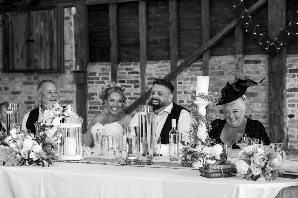A happy wedding reception with five people sitting at a decorated table, laughing and smiling, in a rustic venue with brick walls and string lights.