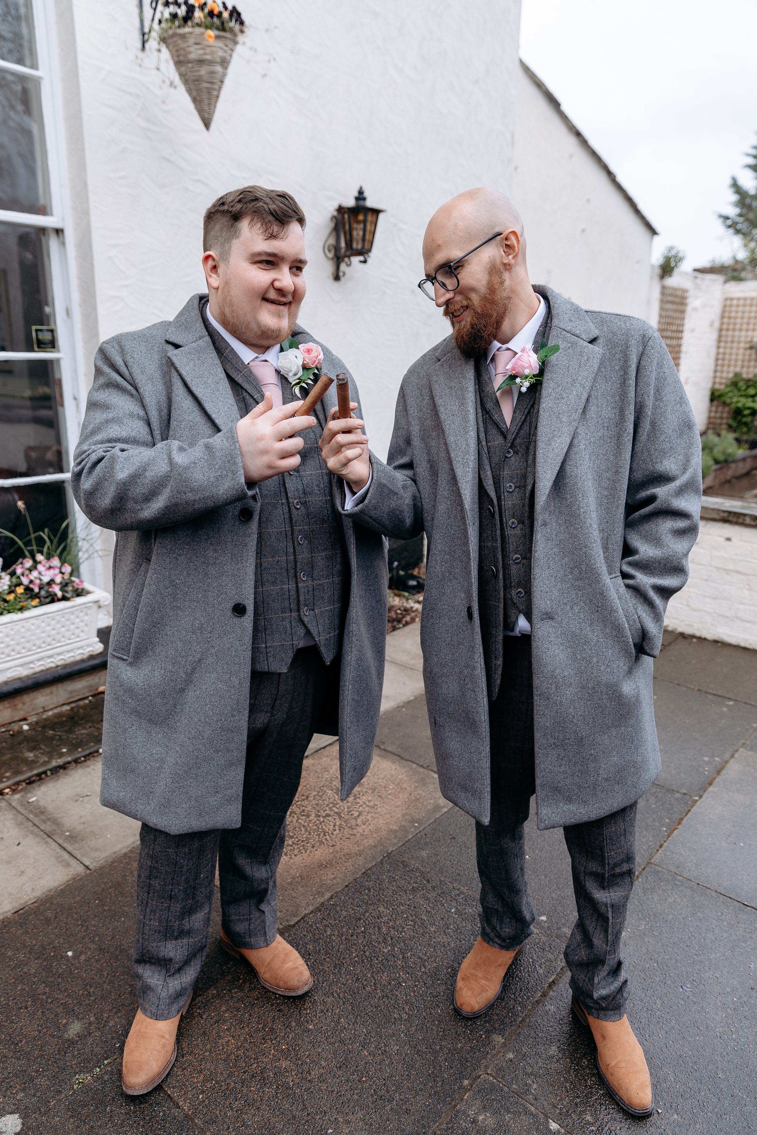 Two men dressed in gray coats, matching patterned pants, and pink ties, holding cigars and toasting each other outside a building with a white wall and hanging flower basket.