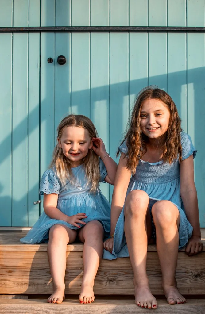 Two young girls in blue dresses sitting on wooden steps in front of a light blue wooden door. They are smiling and the sunlight casts a shadow on the door behind them. Both girls are barefoot.