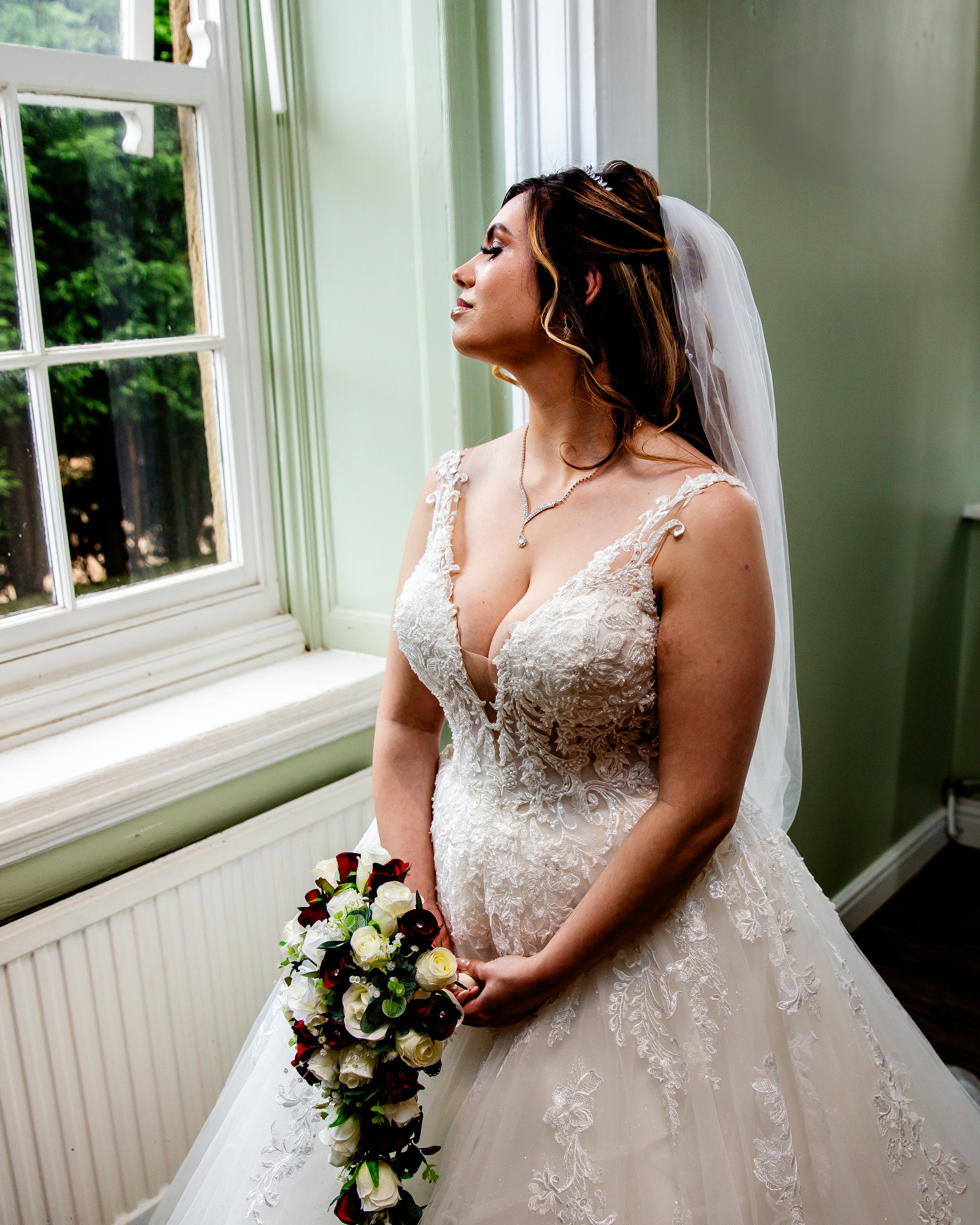Bride in a lace wedding gown standing by a window holding a floral bouquet