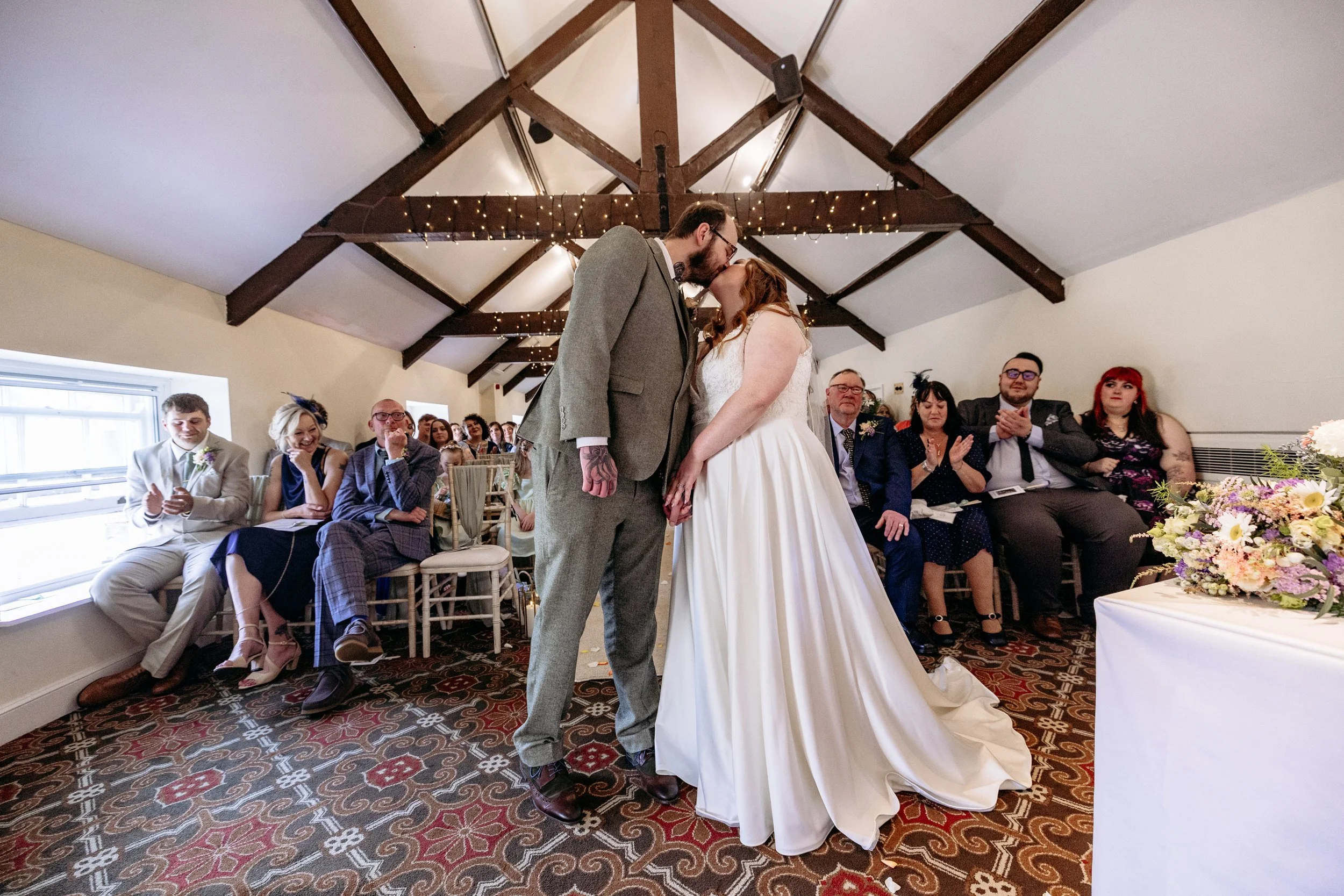 A couple sharing a kiss during their wedding ceremony in a room with wooden beams and fairy lights, with seated guests watching.