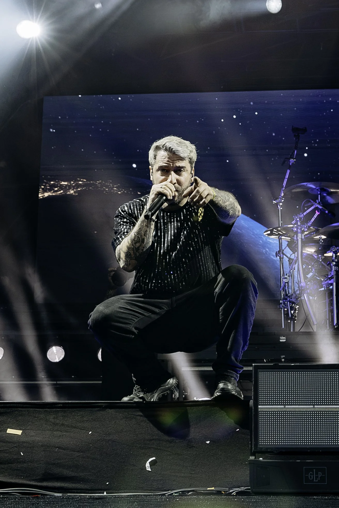 A male singer with tattoos on his arms kneeling on stage, pointing at the camera, holding a microphone, with a digital screen showing earth and space in the background, and stage lights shining down.