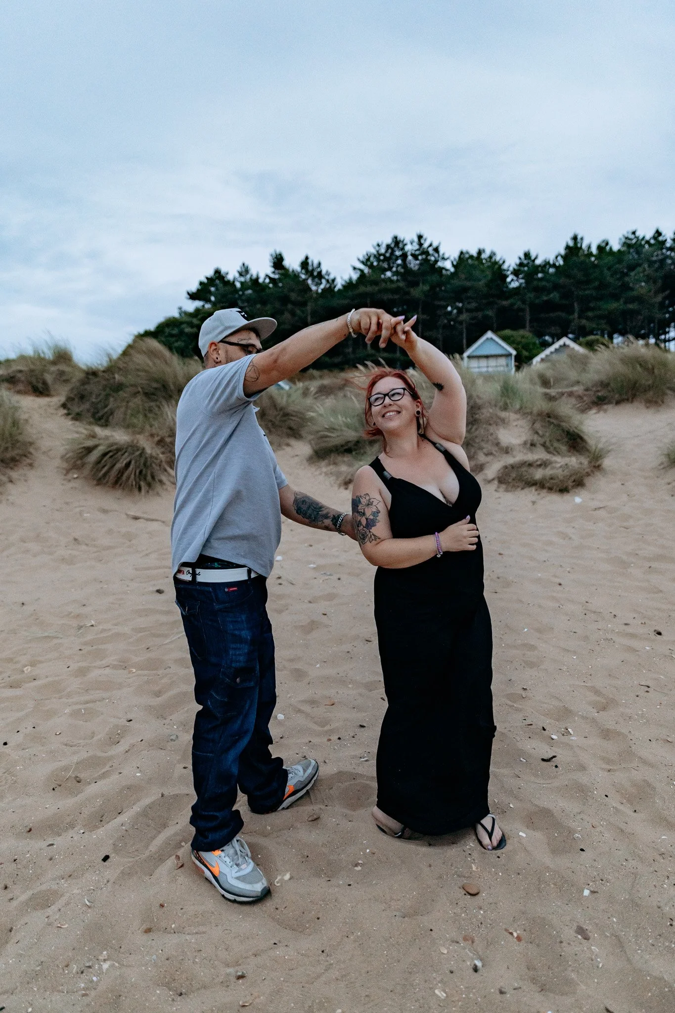 A man and a woman dancing on a sandy beach, with dunes and houses in the background and cloudy sky overhead.
