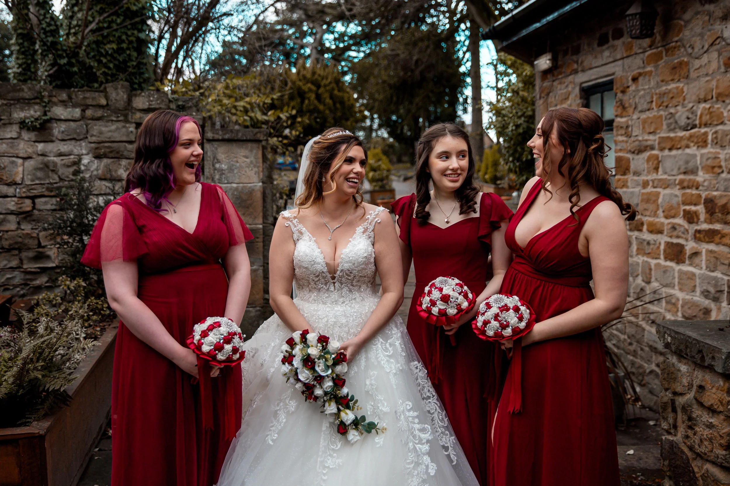 Bride and four bridesmaids standing outdoors in front of a stone wall, smiling and holding bouquets, at a wedding.