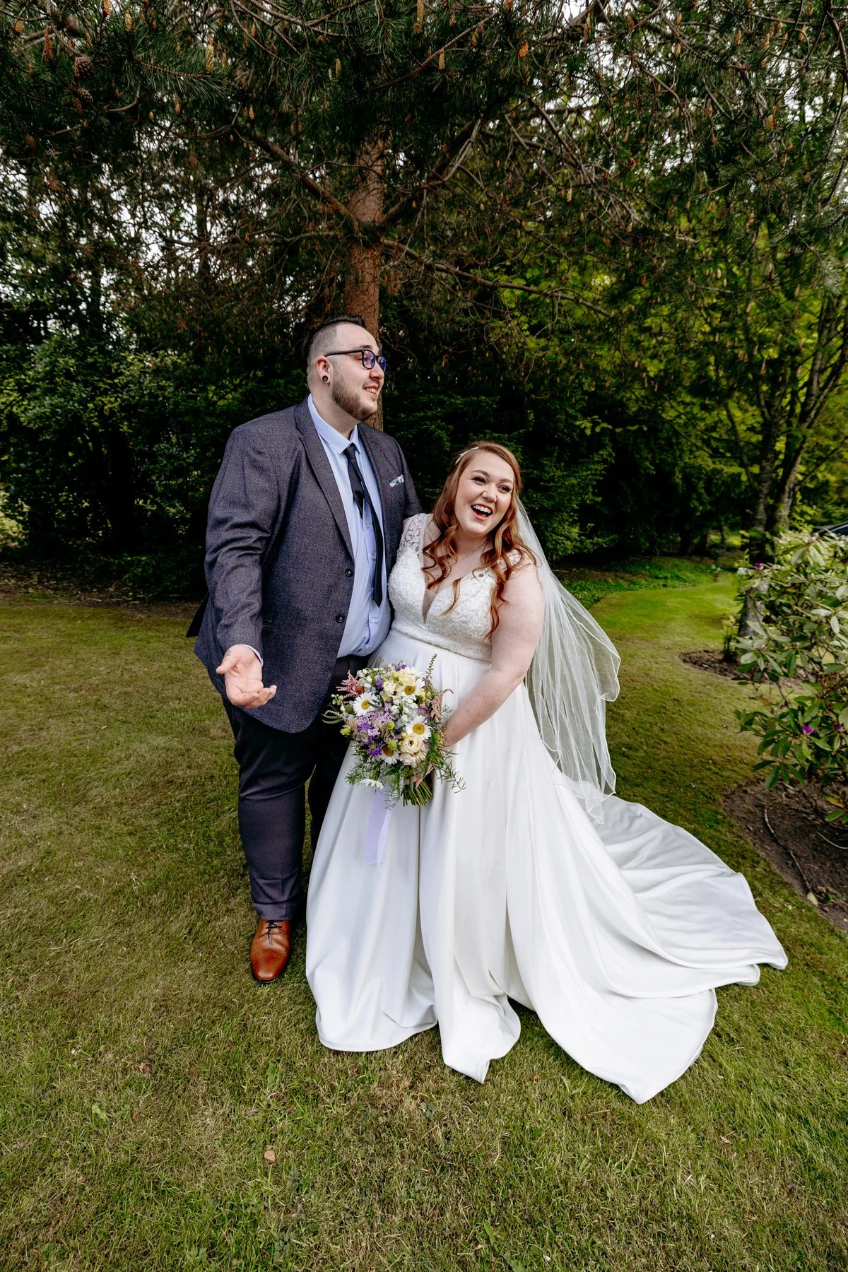 A newlywed couple standing on a grassy lawn with trees in the background, smiling and laughing. The bride is wearing a white wedding gown and holding a bouquet of flowers, while the groom is dressed in a dark gray suit with a blue shirt and tie.