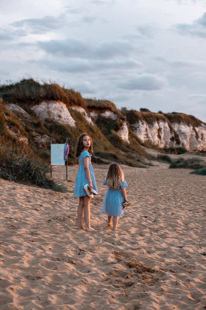 Two girls in blue dresses walking barefoot on a sandy beach with cliffs in the background.