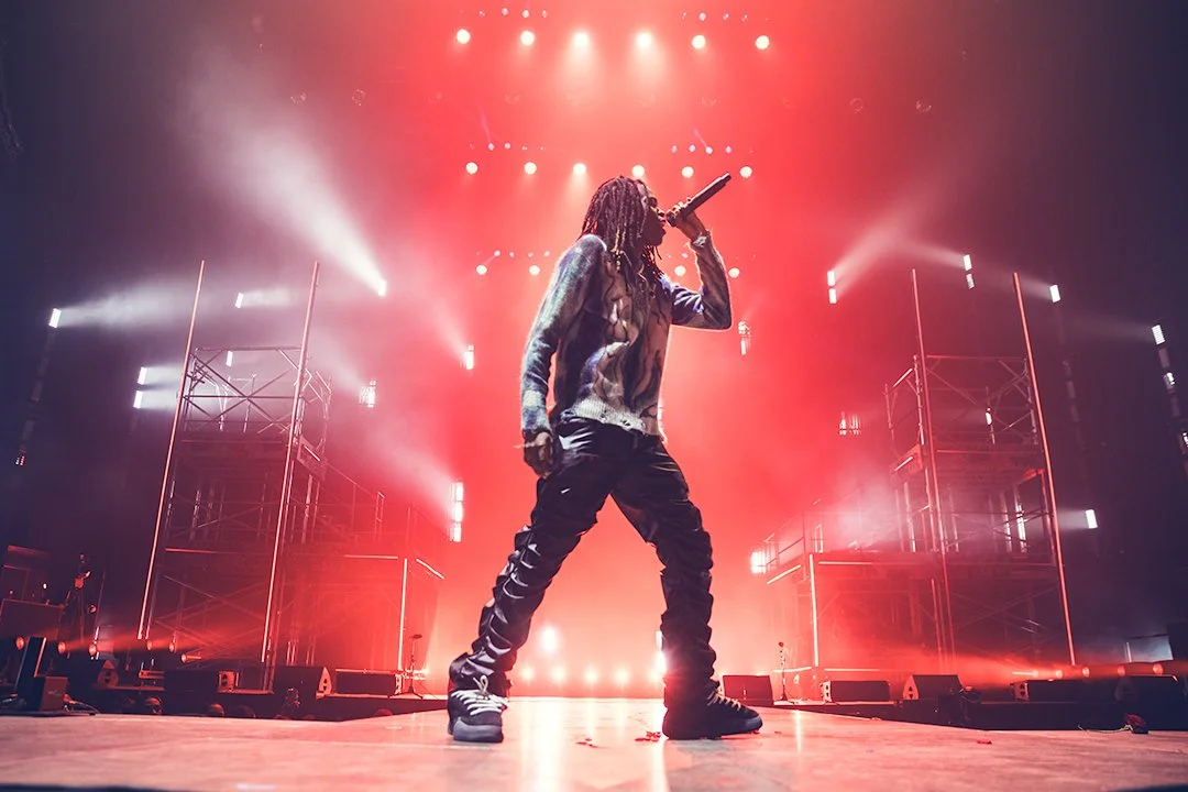 A performer with dreadlocks singing into a microphone on stage with red and white lights and scaffolding in the background.