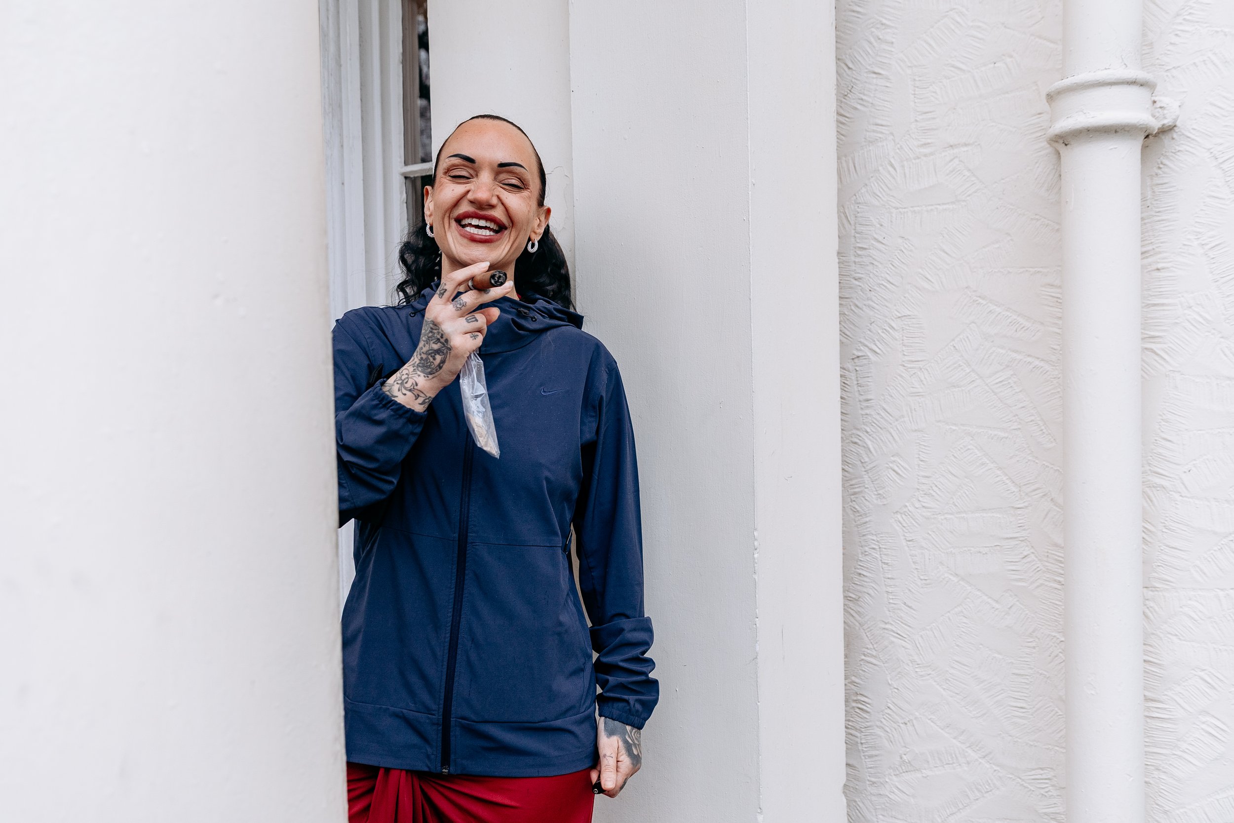 A woman standing in a doorway, smiling and holding a snack, dressed in a blue jacket and red skirt, with tattoos on her hand and neck.