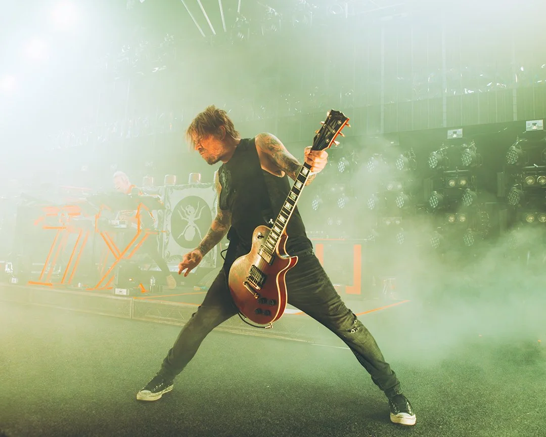 Musician playing electric guitar on stage surrounded by yellow-green stage fog, with DJ equipment and stage lights in background.