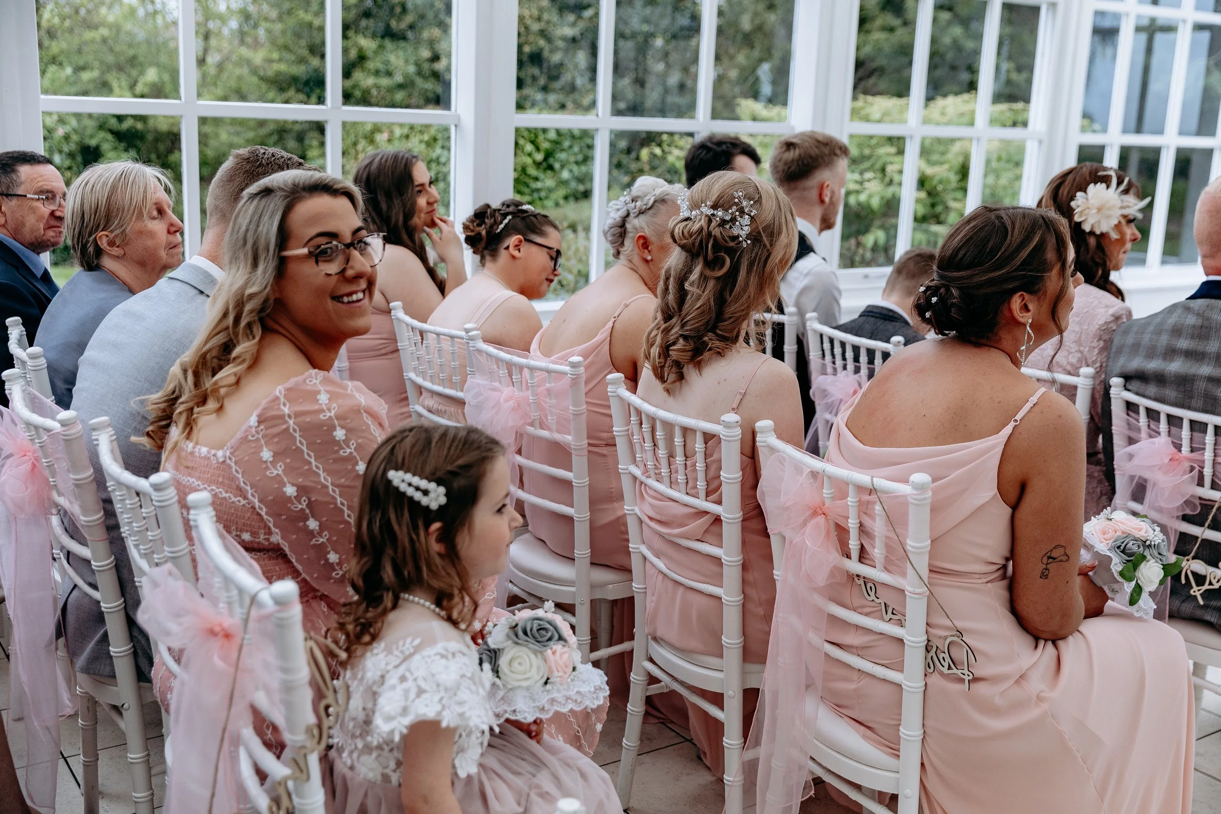 Group of women and children sitting in a bright, floral greenhouse or conservatory, dressed in pink and light-colored formal attire for a wedding or similar event, with outdoor greenery visible through the windows.