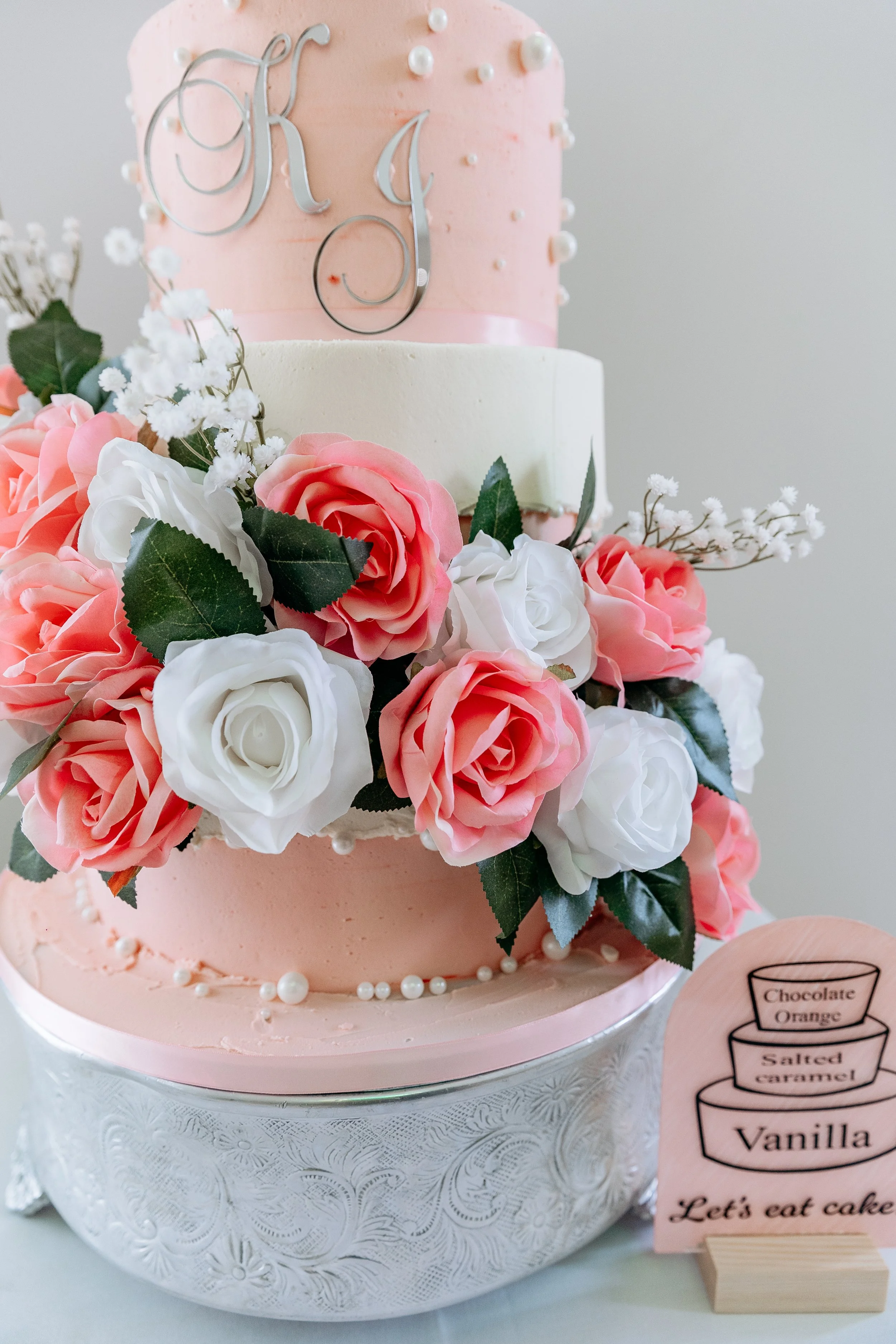 Three-tiered wedding cake decorated with pink and white flowers, pearls, and metallic initials on the top tier, placed in an ornate silver cake stand, with a wooden sign listing cake flavors.