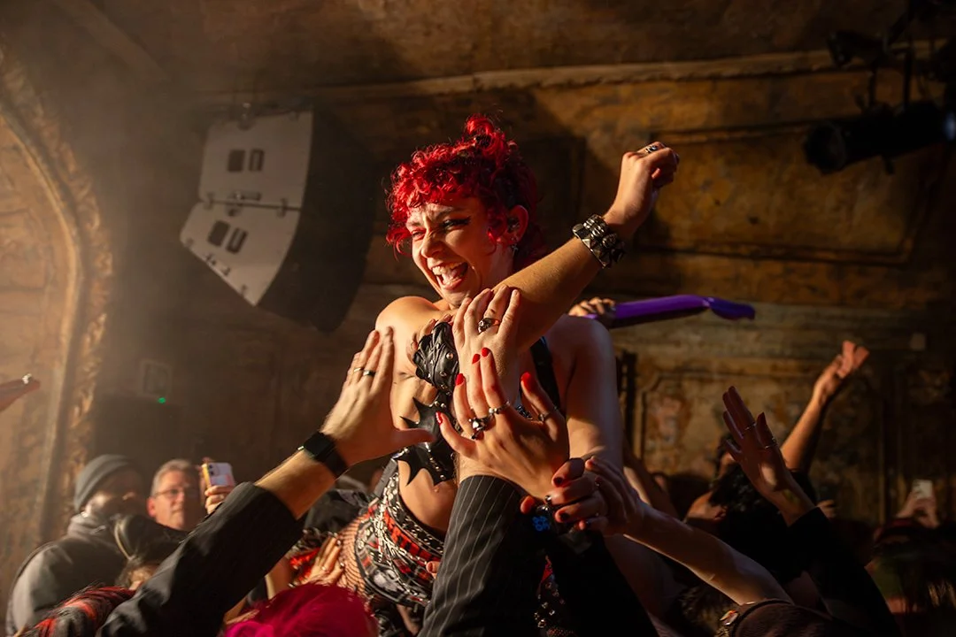 A woman with bright red, curly hair dancing and smiling in a crowded, lively club scene. She is surrounded by people reaching out to her, with some holding up their phones.