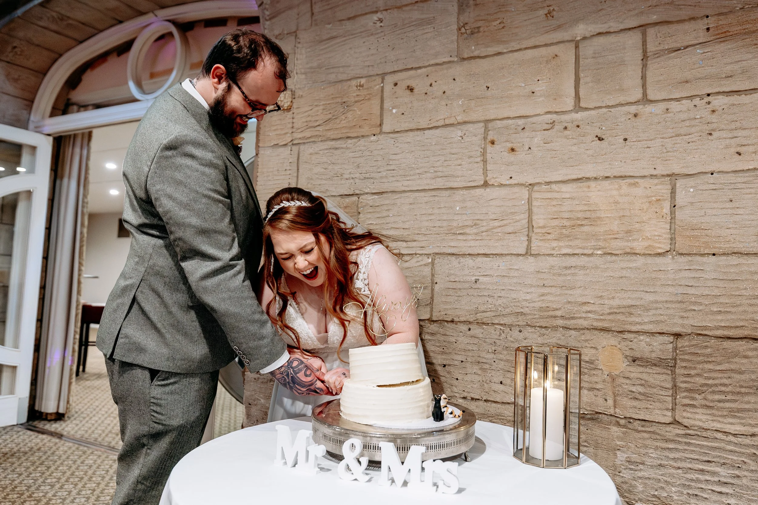 A bride and groom cutting their wedding cake together, smiling and laughing, at a rustic wedding reception with beige brick walls and a white table with wedding signs and candle centerpiece.