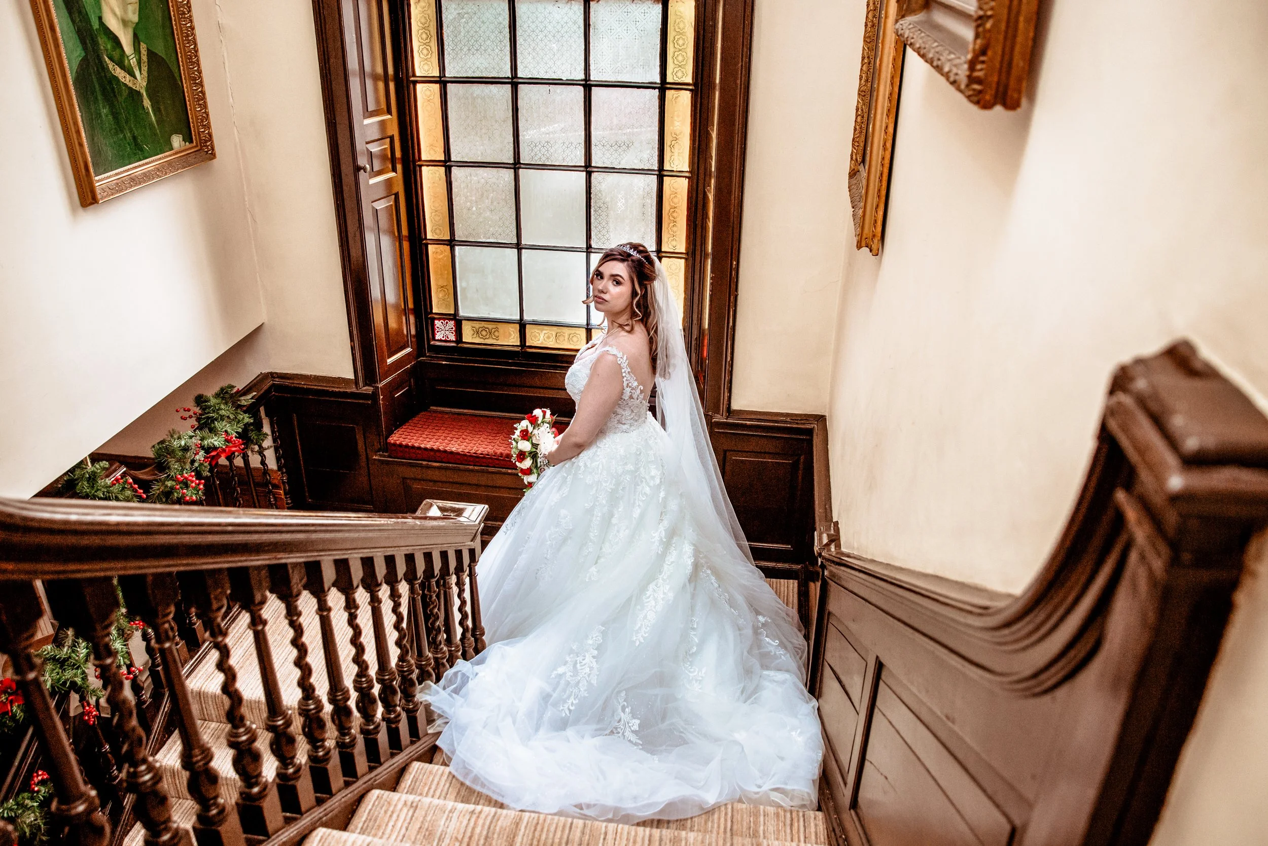 Bridal gown with lace details, carrying a bouquet of roses, standing on a staircase inside a vintage building with stained glass windows and wooden paneling.