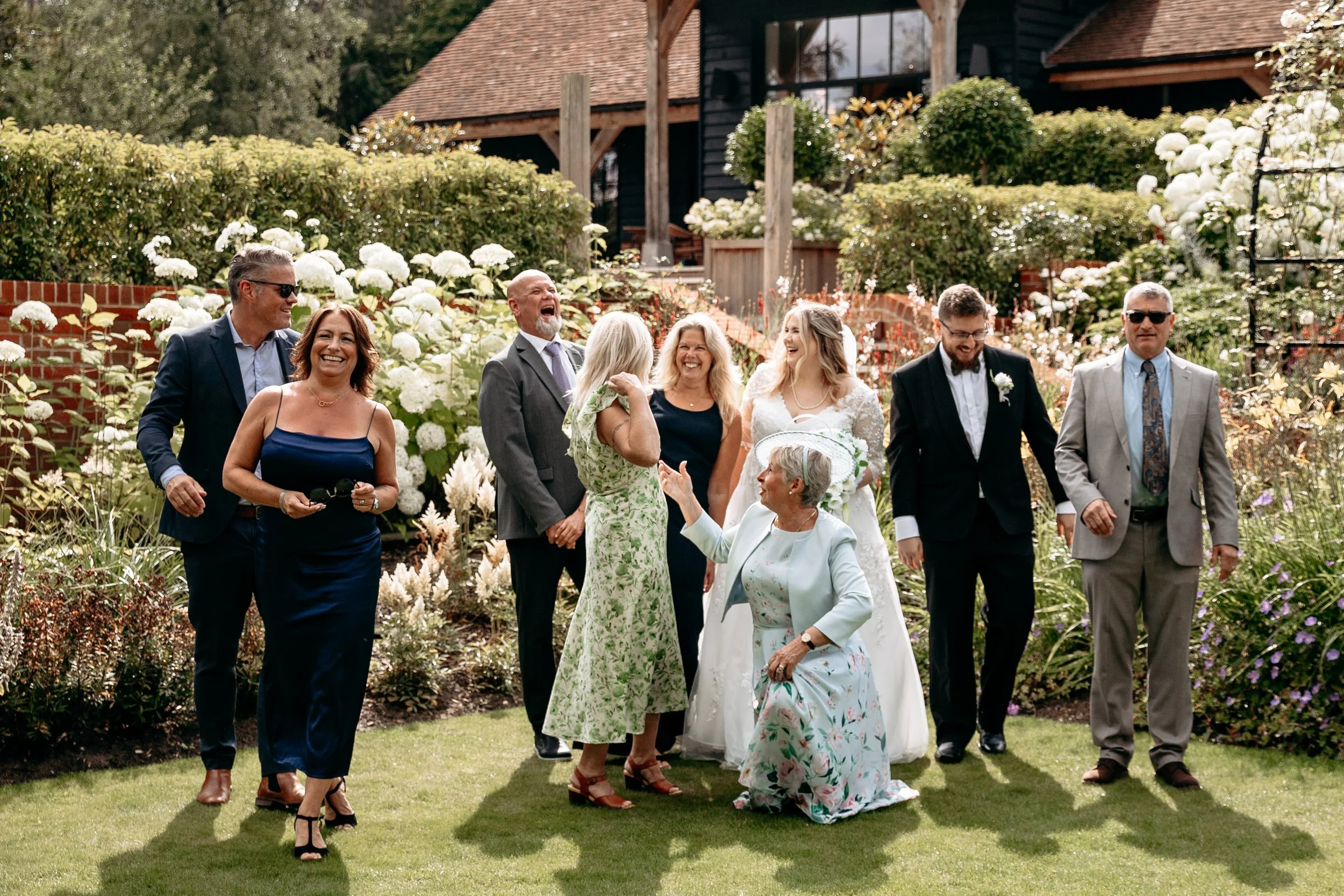Group of people laughing together in a garden, surrounded by greenery and white flowers, with a rustic building in the background.