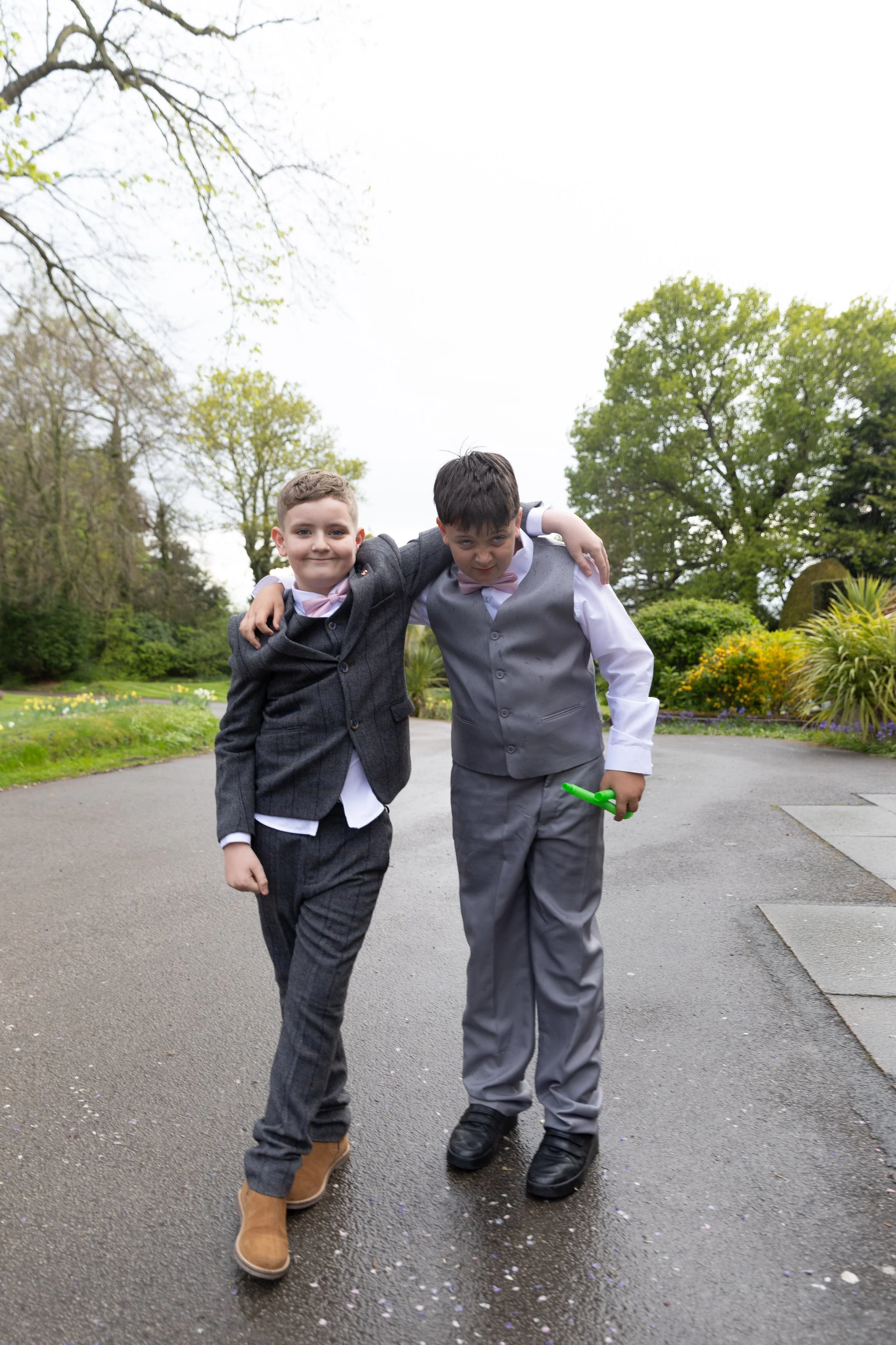 Two young boys in suits walking together outdoors on a paved path, with one arm around each other's shoulders, smiling and looking at the camera, with trees and bushes in the background.