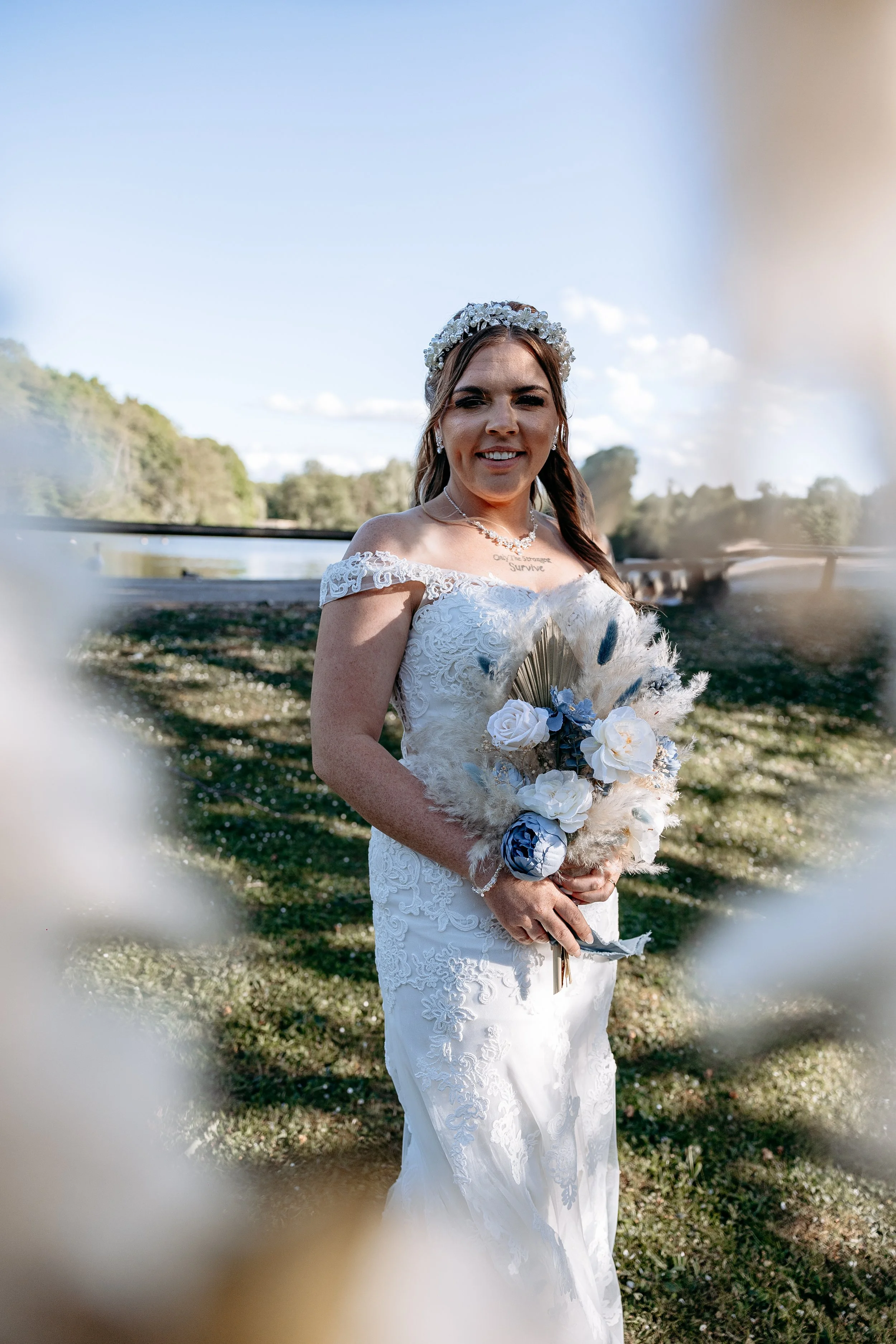 A bride in a white lace wedding dress holding a bouquet of flowers and pampas grass, standing outdoors on a bright day with trees and a blue sky in the background.