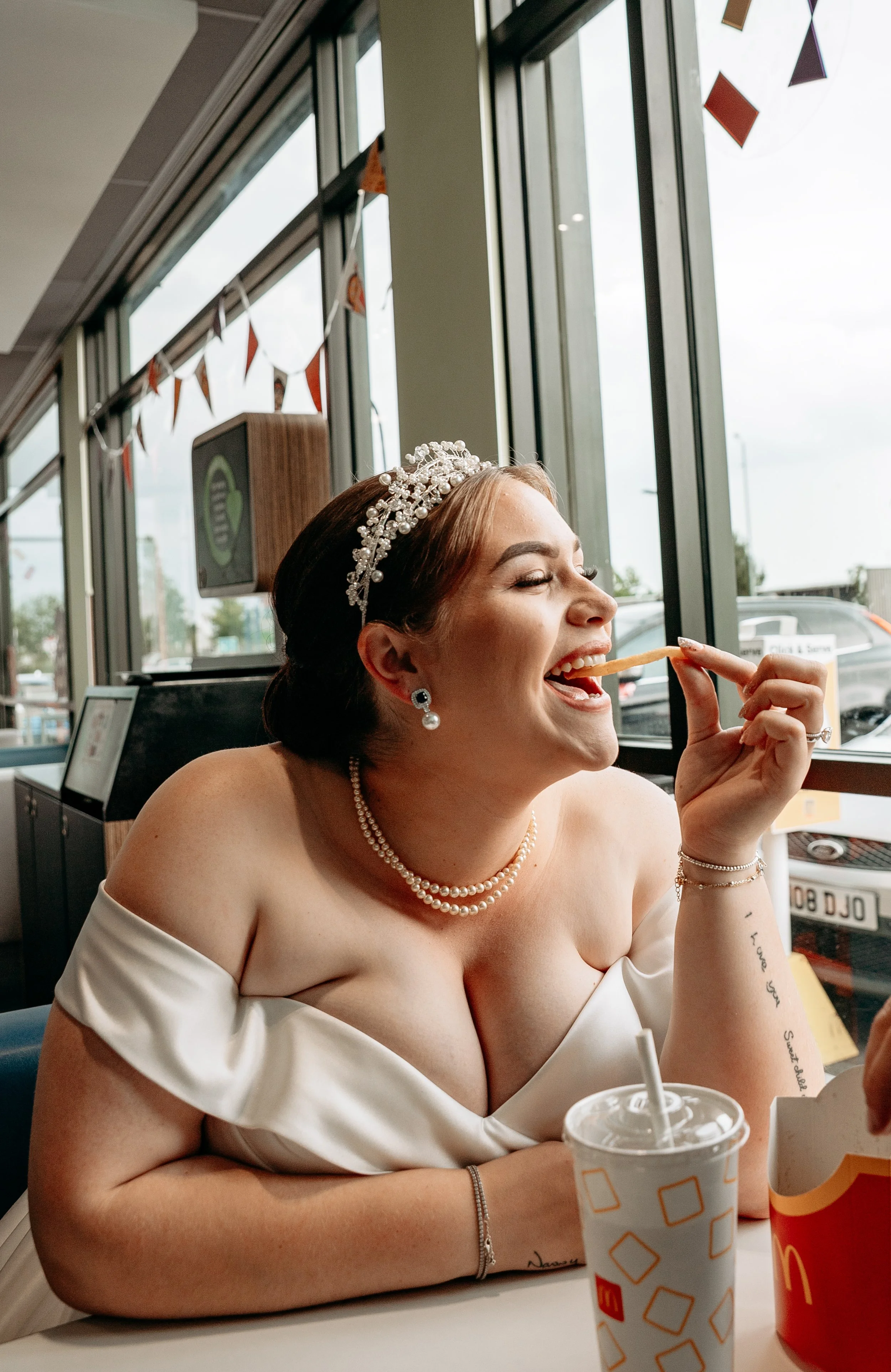 A woman in a wedding dress with pearl jewelry and an ornate headpiece enjoying French fries inside a fast food restaurant.