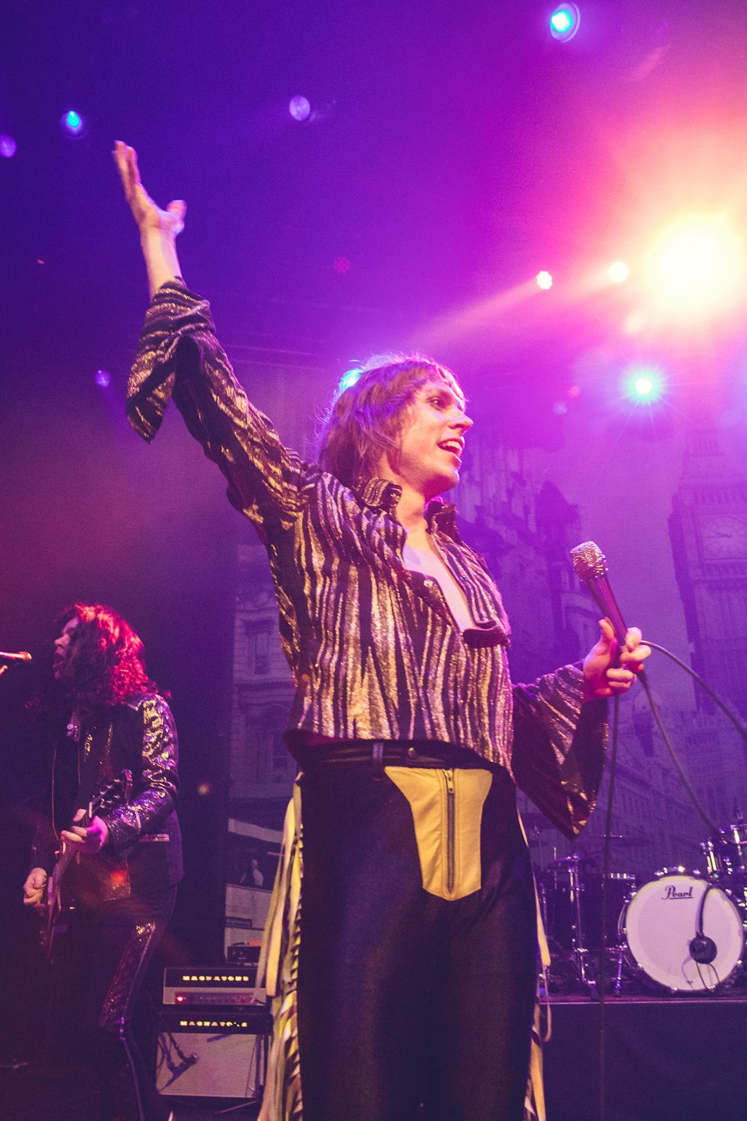 A performer wearing a metallic, striped shirt and black pants is on stage with a microphone, raising one hand. Another musician is playing guitar in the background. The stage is lit in purple and orange lights, with a drum set also visible.