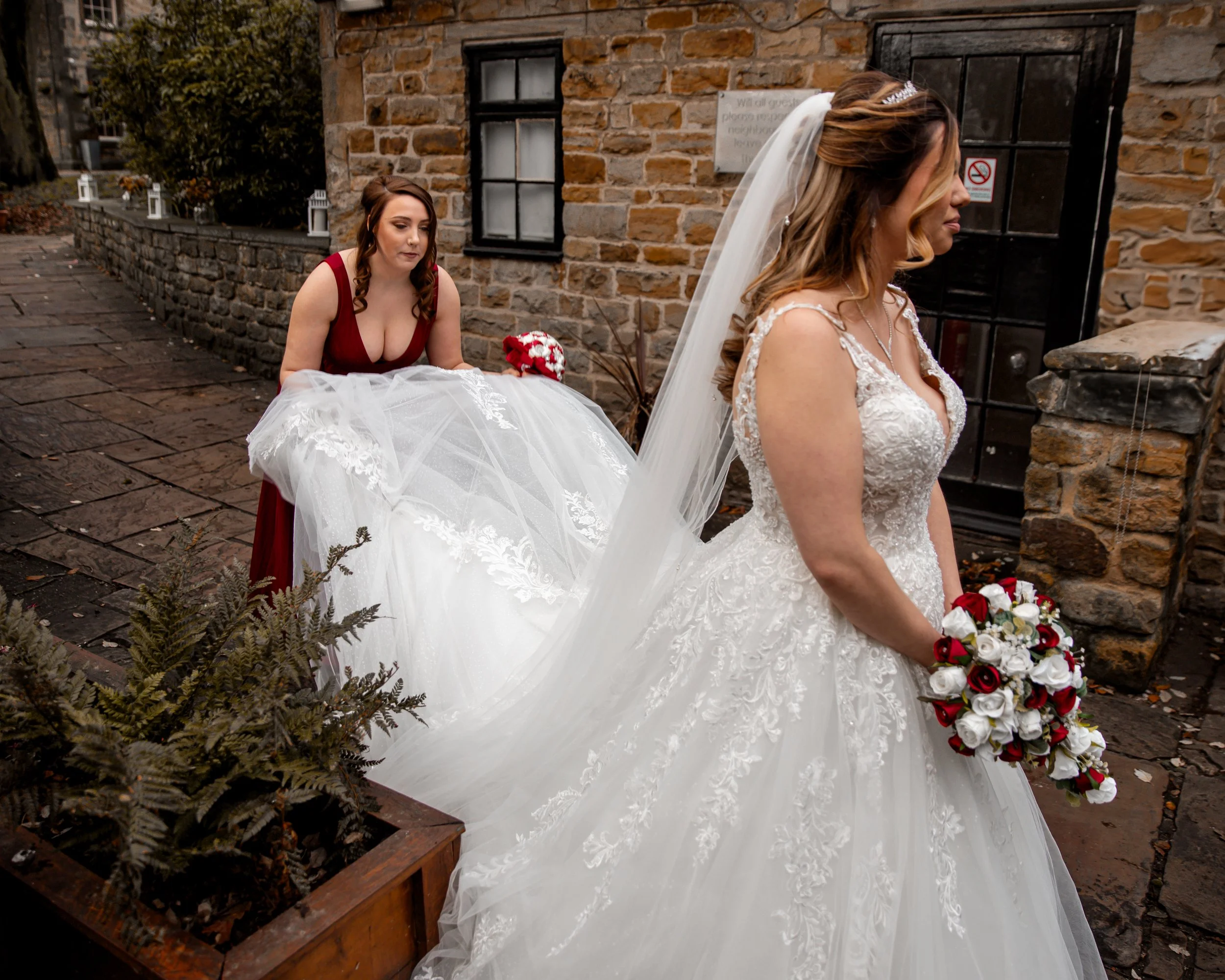 Bride in a white wedding gown holding a bouquet of red and white roses, with a bridesmaid in a red dress assisting her outside a brick building.