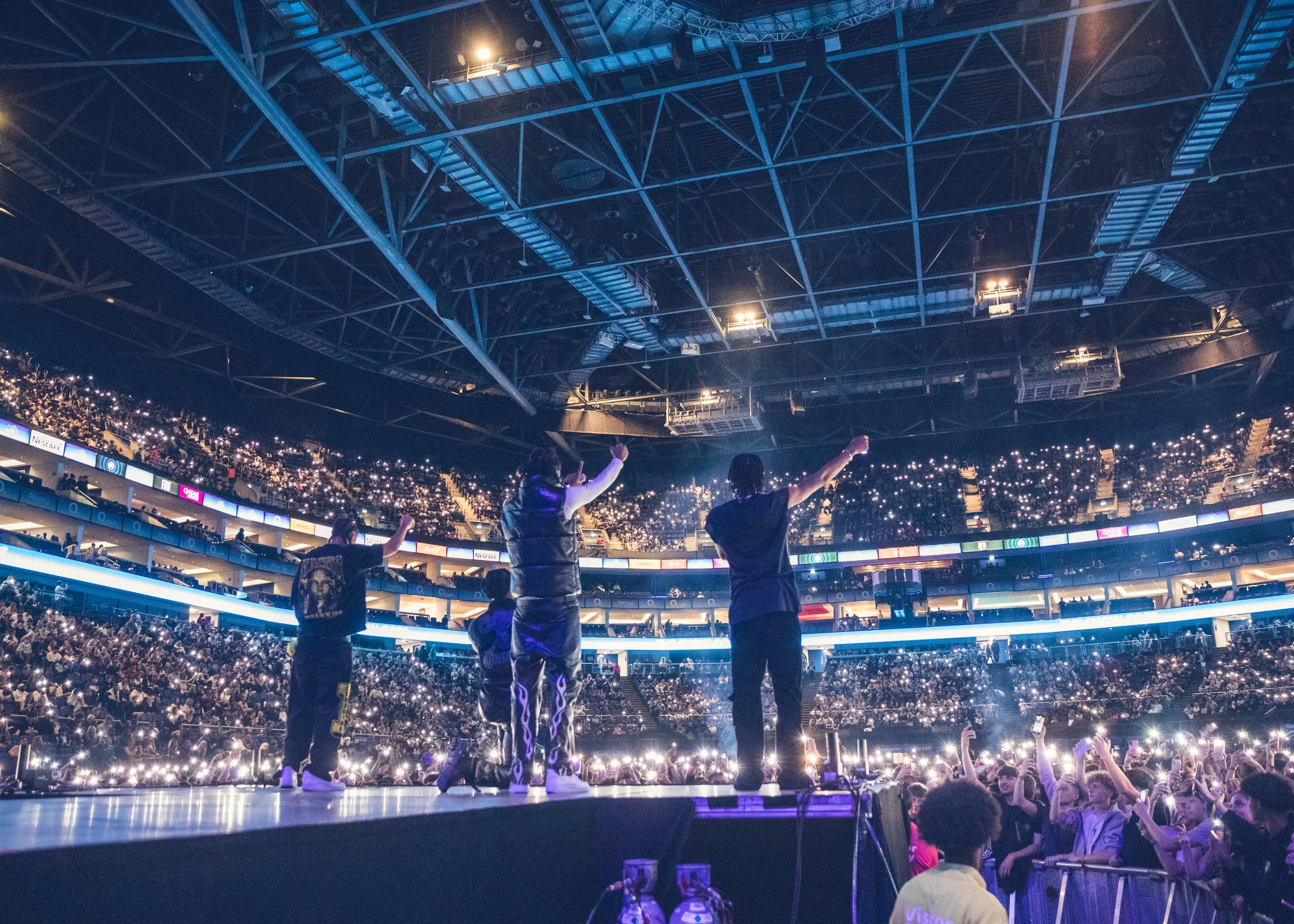 Three performers on stage in a large arena, facing a cheering crowd with lights from audience members' phones illuminating the scene; the venue is packed with spectators.