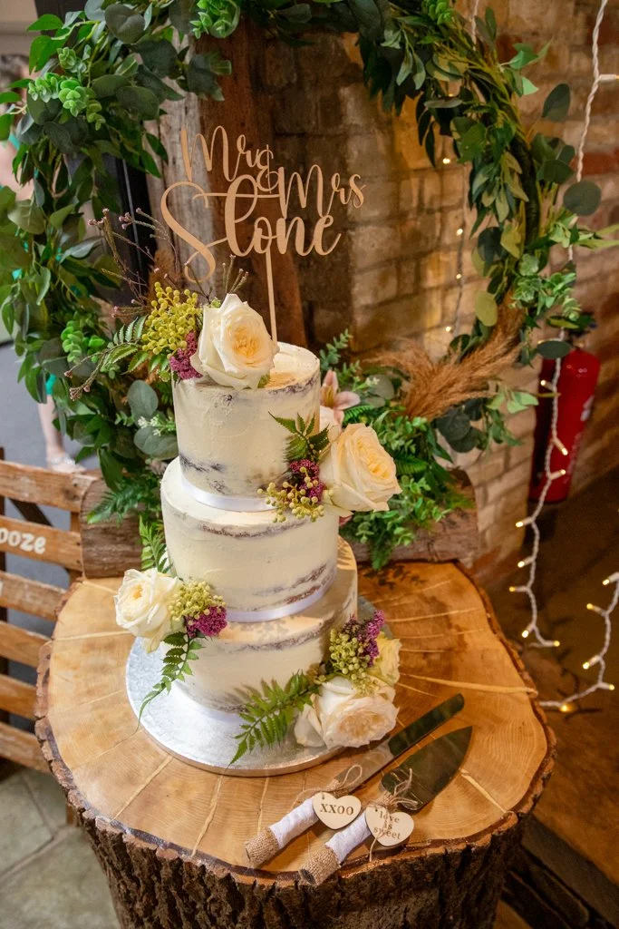 Three-tiered wedding cake decorated with white roses and assorted greenery, with a wooden 'Mrs. & Mrs. Cone' cake topper on a rustic wooden table.