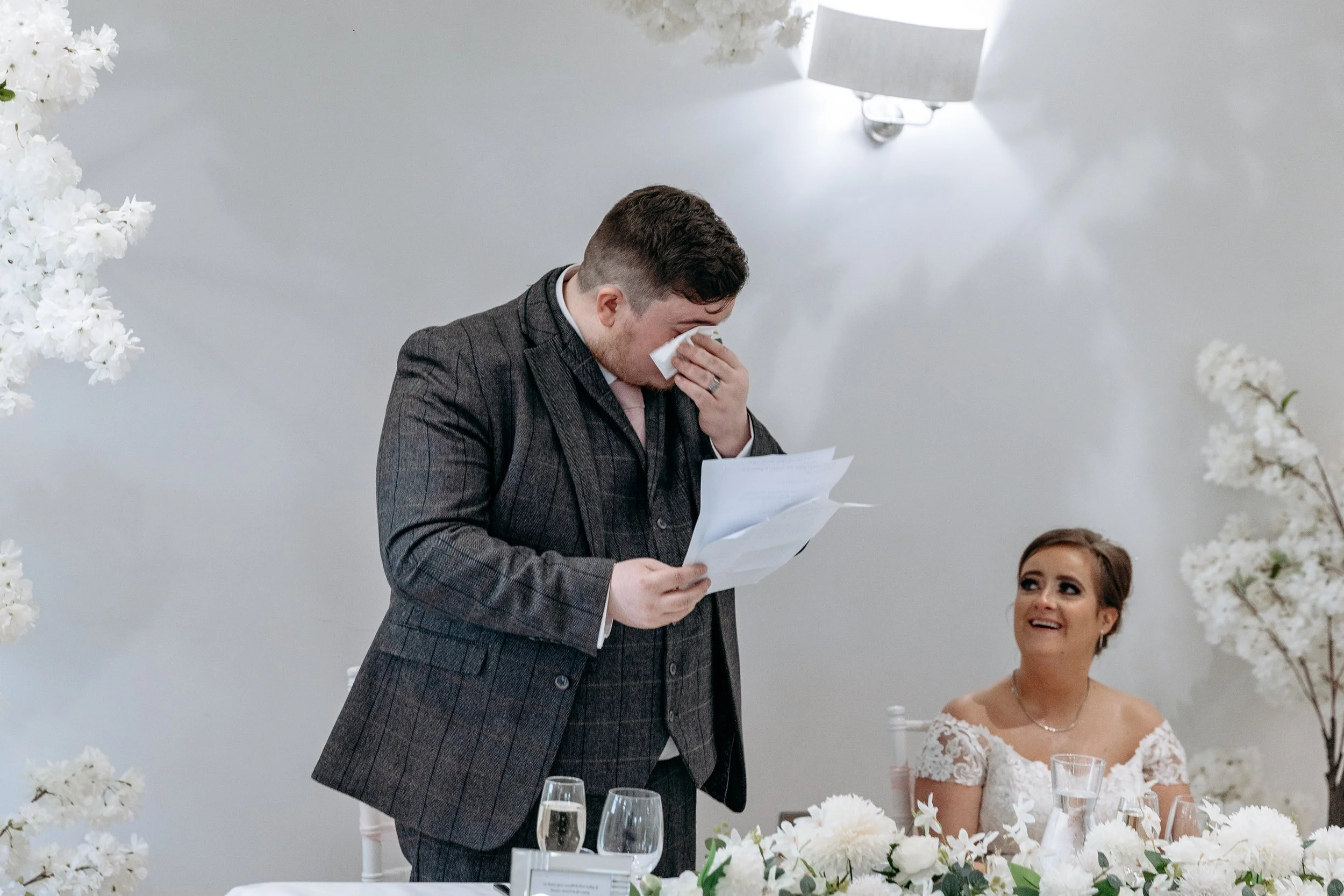 A man in a dark gray suit reading from papers while holding a tissue to his face, standing at a wedding reception table. A woman in a white lace wedding dress is sitting and looking up at him with a smile. The table is decorated with white flowers an