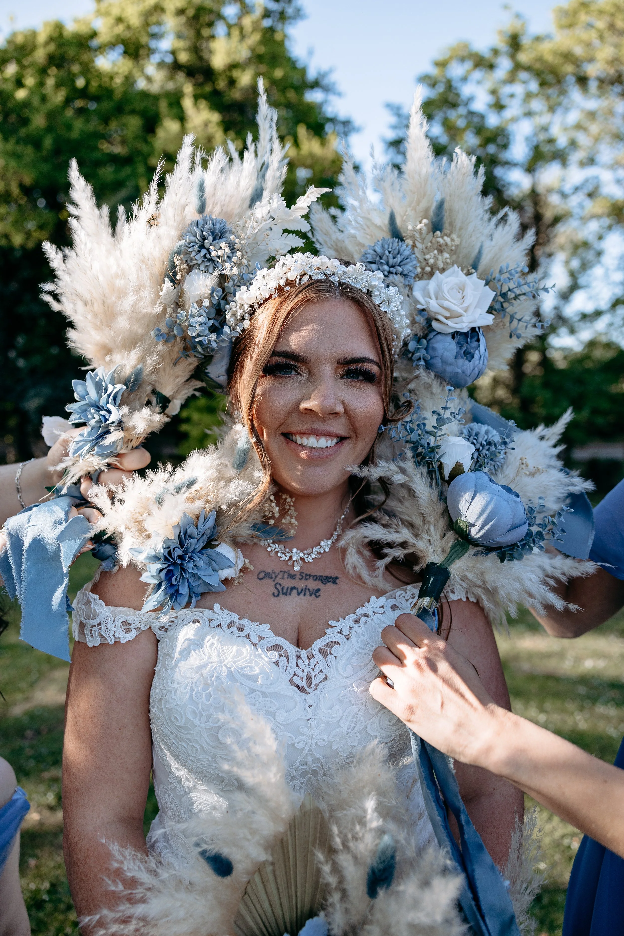 A smiling bride wearing a white lace wedding dress with a floral tattoo on her chest, holding a large bouquet of white and blue flowers, with an elaborate headdress made of pampas grass, flowers, and ribbons